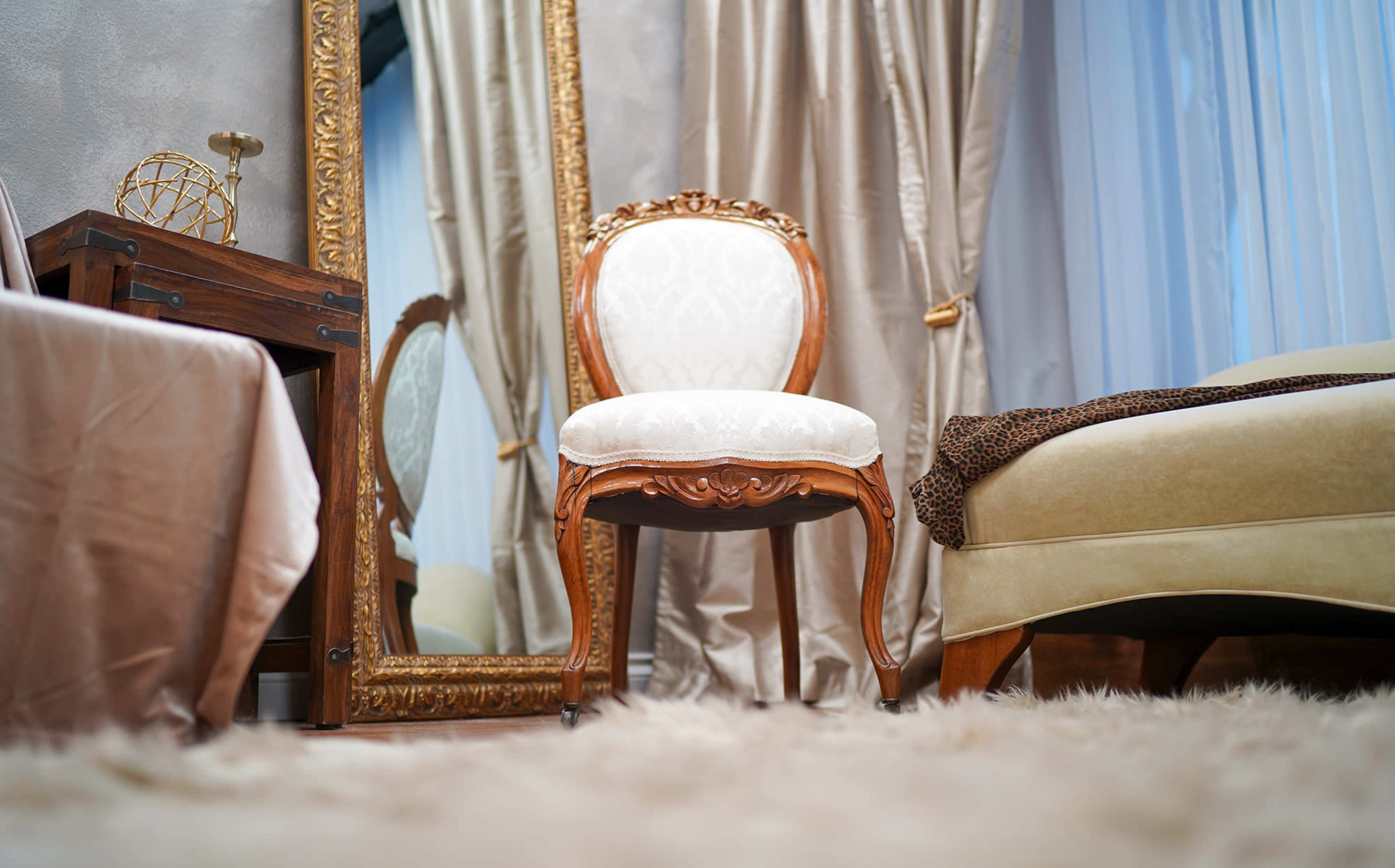 The image features an ornate wooden chair facing a large mirror in a softly lit room with beige and gold decor.
