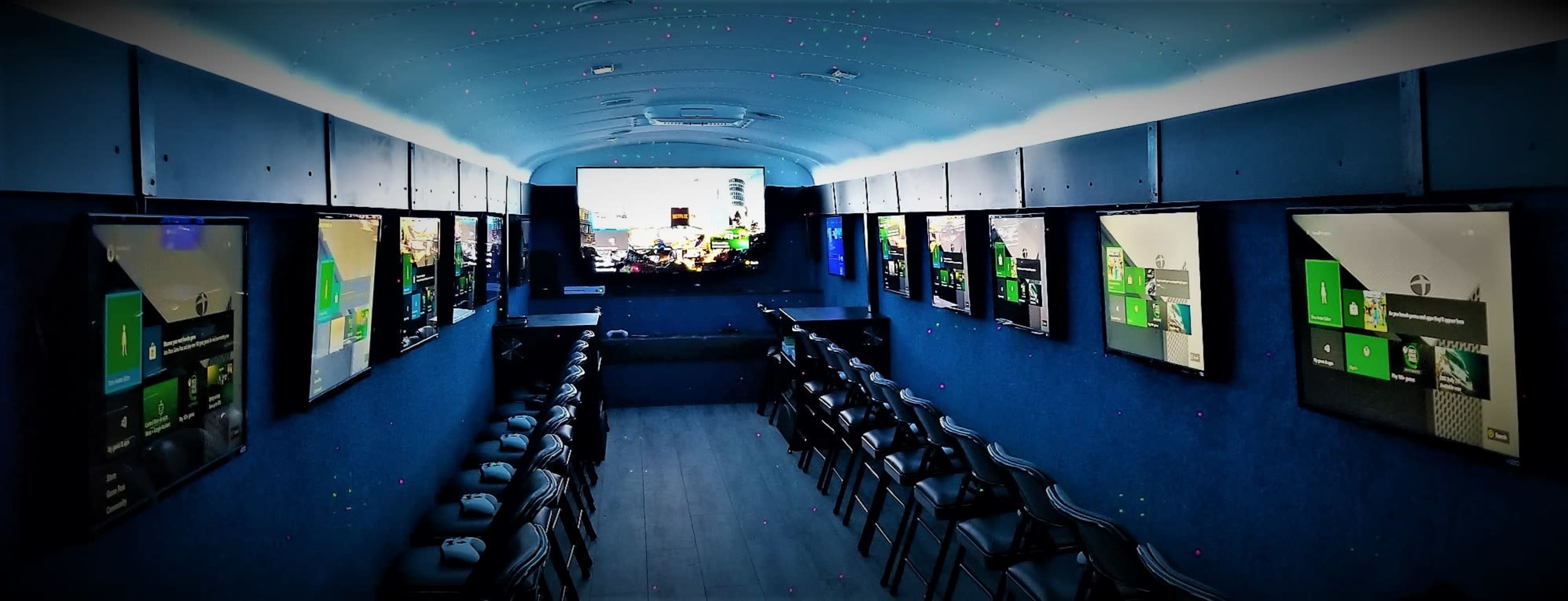 The image shows an interior of a mobile gaming setup featuring multiple screens mounted on the walls, with rows of chairs and gaming consoles arranged for players.