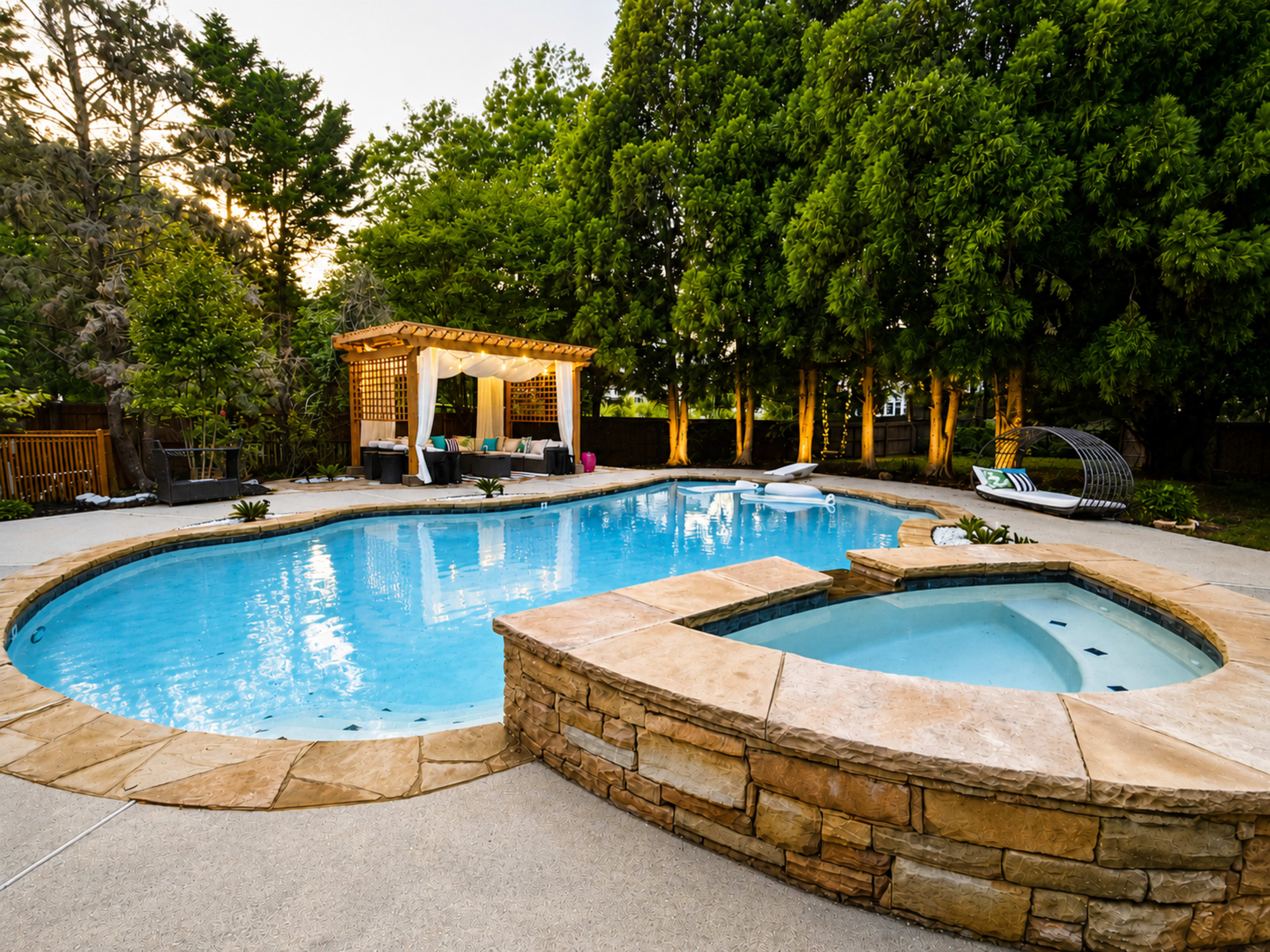 A curved swimming pool with a hot tub is surrounded by landscaped greenery and a pergola in a backyard setting.