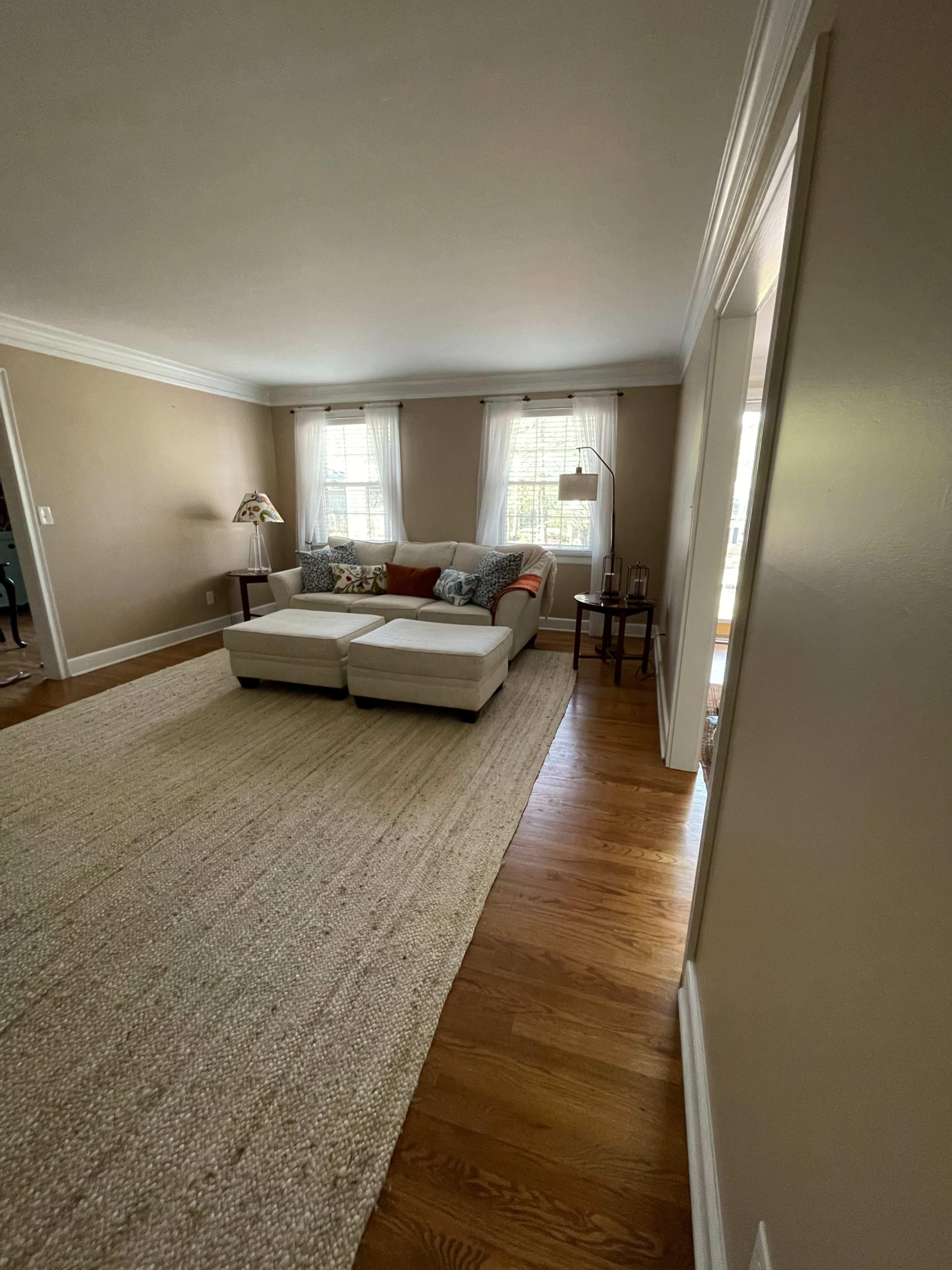 A light-filled living room features a white sectional sofa, a woven area rug, and windows draped with sheer curtains.