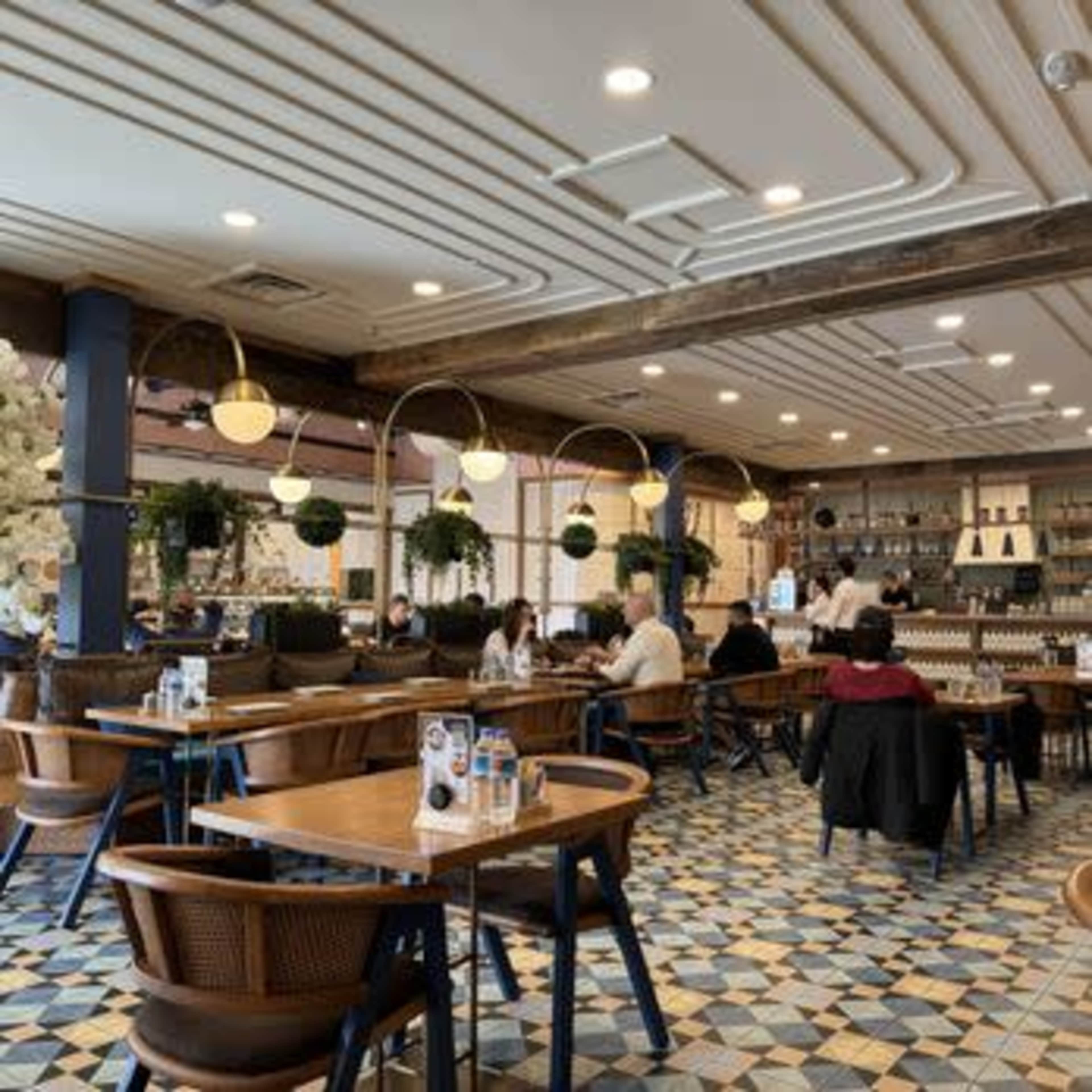 The image shows a modern restaurant interior with wooden tables, chairs, and a patterned tiled floor, featuring plants and a bar area in the background.