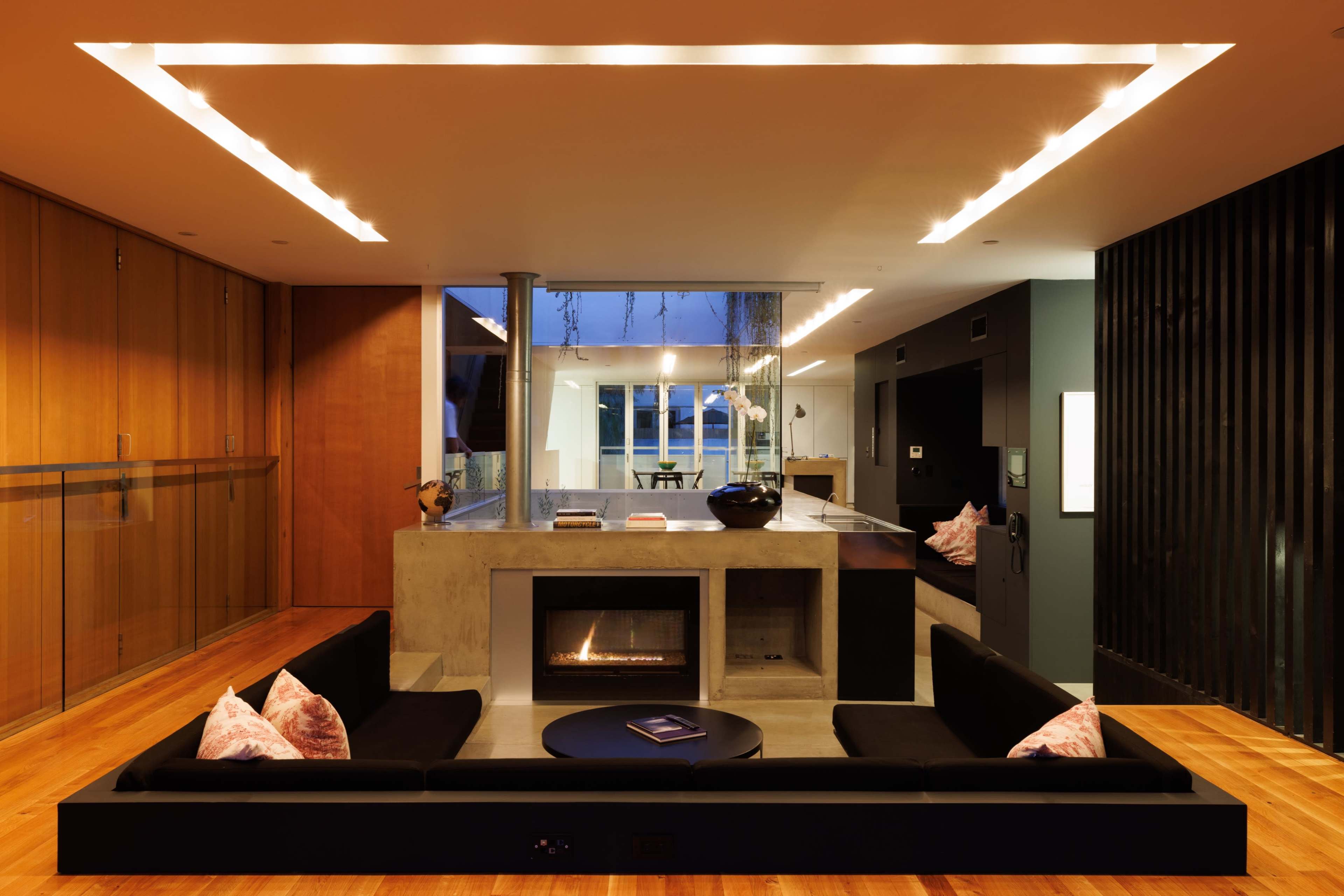 The interior features a modern living space with a central fireplace, black sofas, and large windows that reveal an evening view.