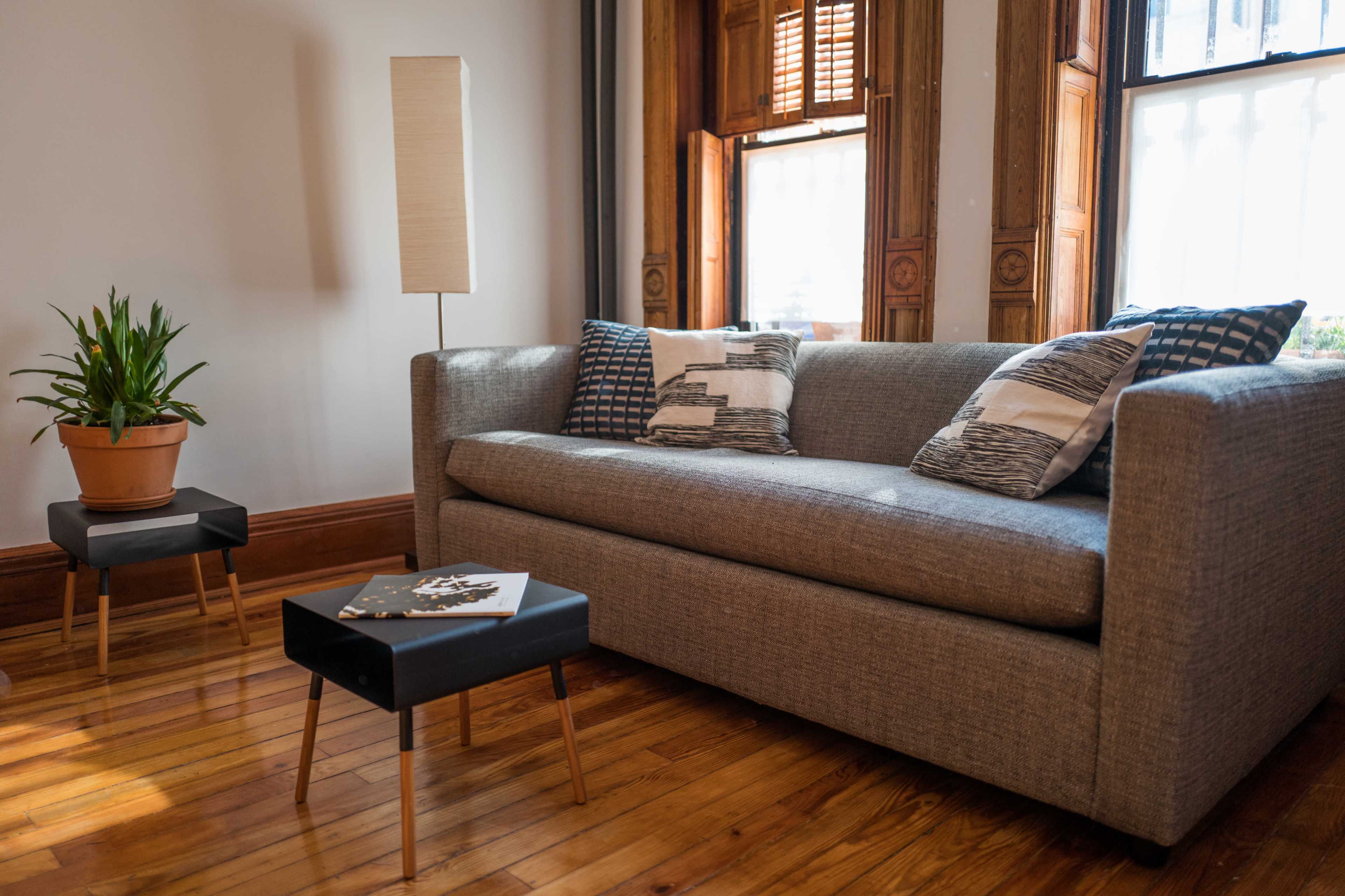 A gray sofa with patterned cushions placed beside a potted plant and a small black side table in a well-lit room with wooden flooring.