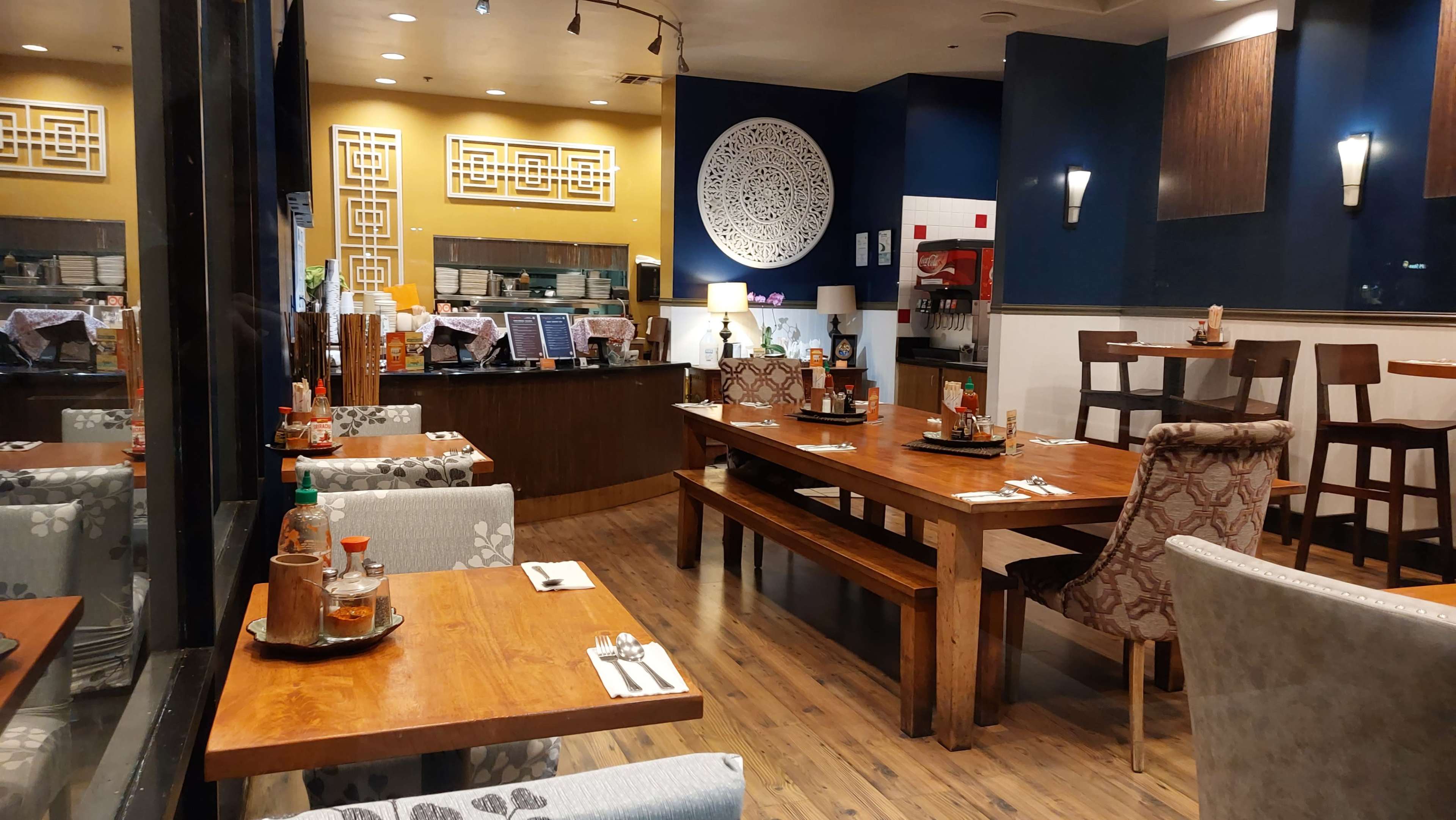The image shows a modern restaurant interior featuring wooden tables and chairs, with a decorative wall design and an area for food service in the background.