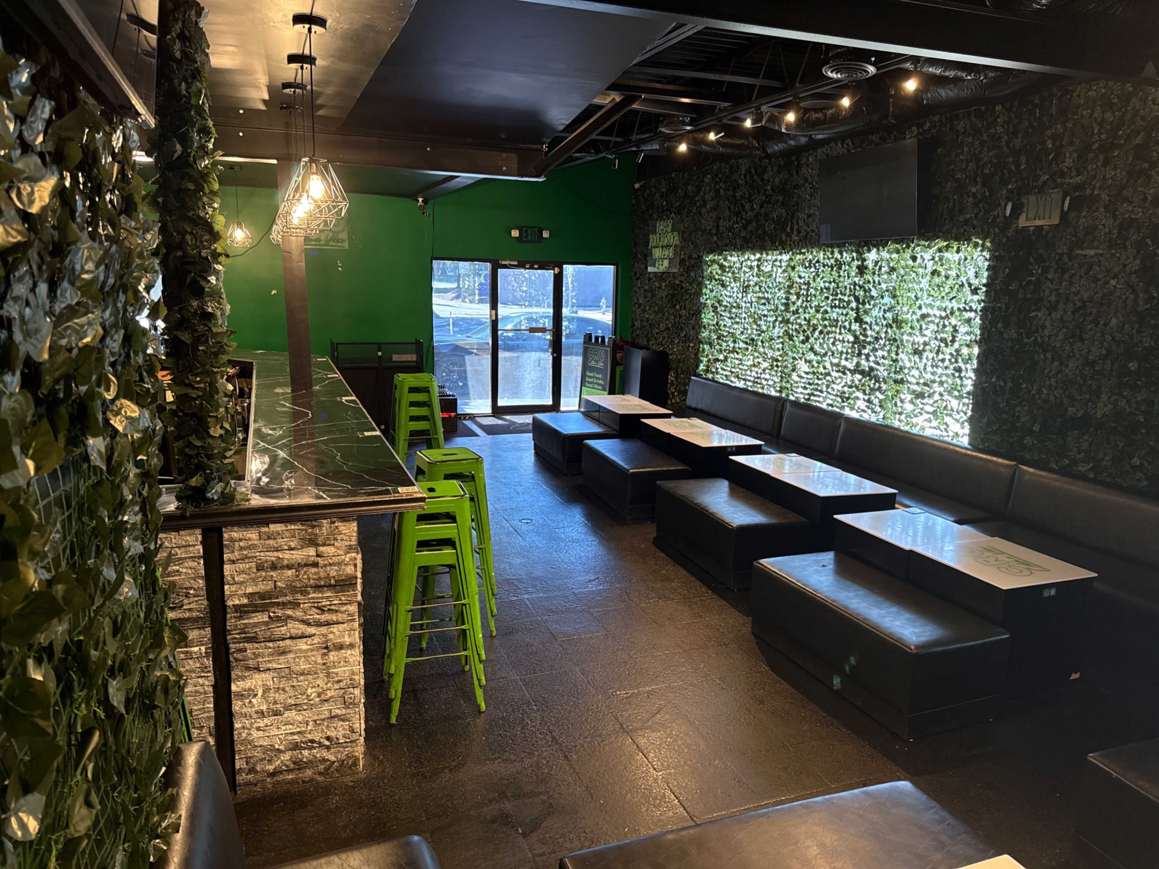 The image shows a bar area with green walls, a dark seating area, and large windows allowing natural light to enter.