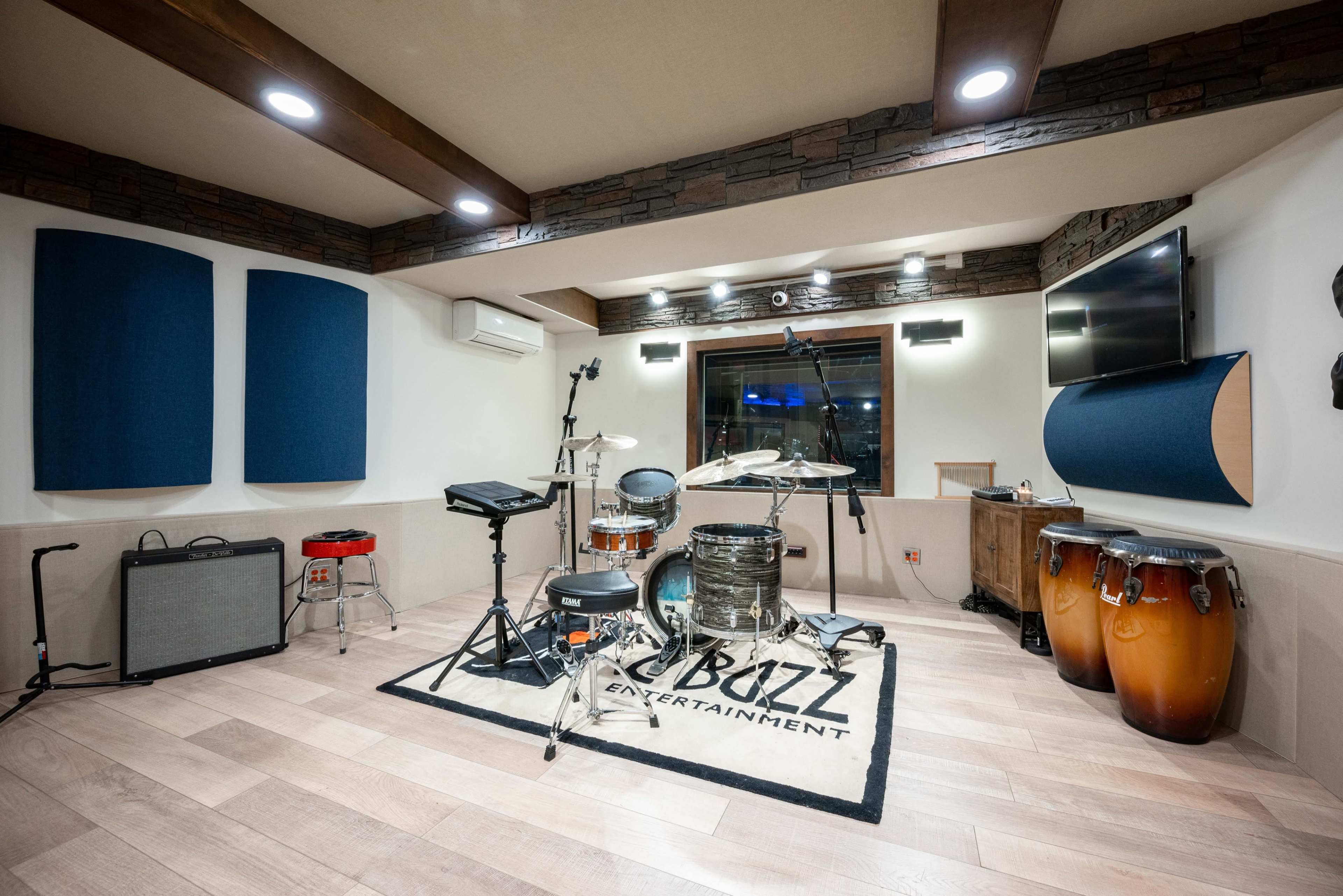 A well-equipped music studio featuring a drum set, conga drums, and various electronic music instruments arranged on a wooden floor with acoustic panels on the walls.