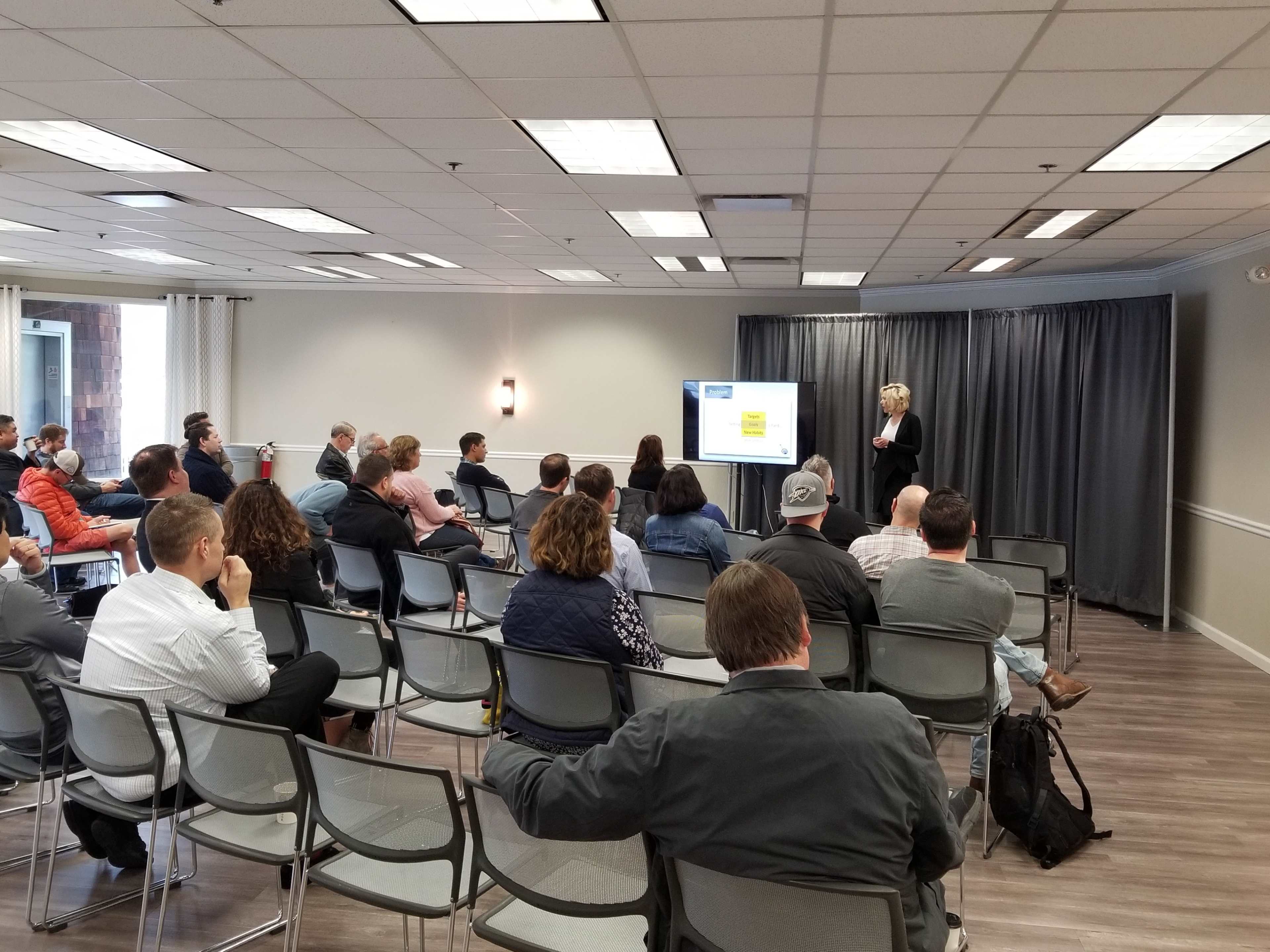 A presenter stands in front of an audience in a conference room, with a PowerPoint slide displayed on a screen.