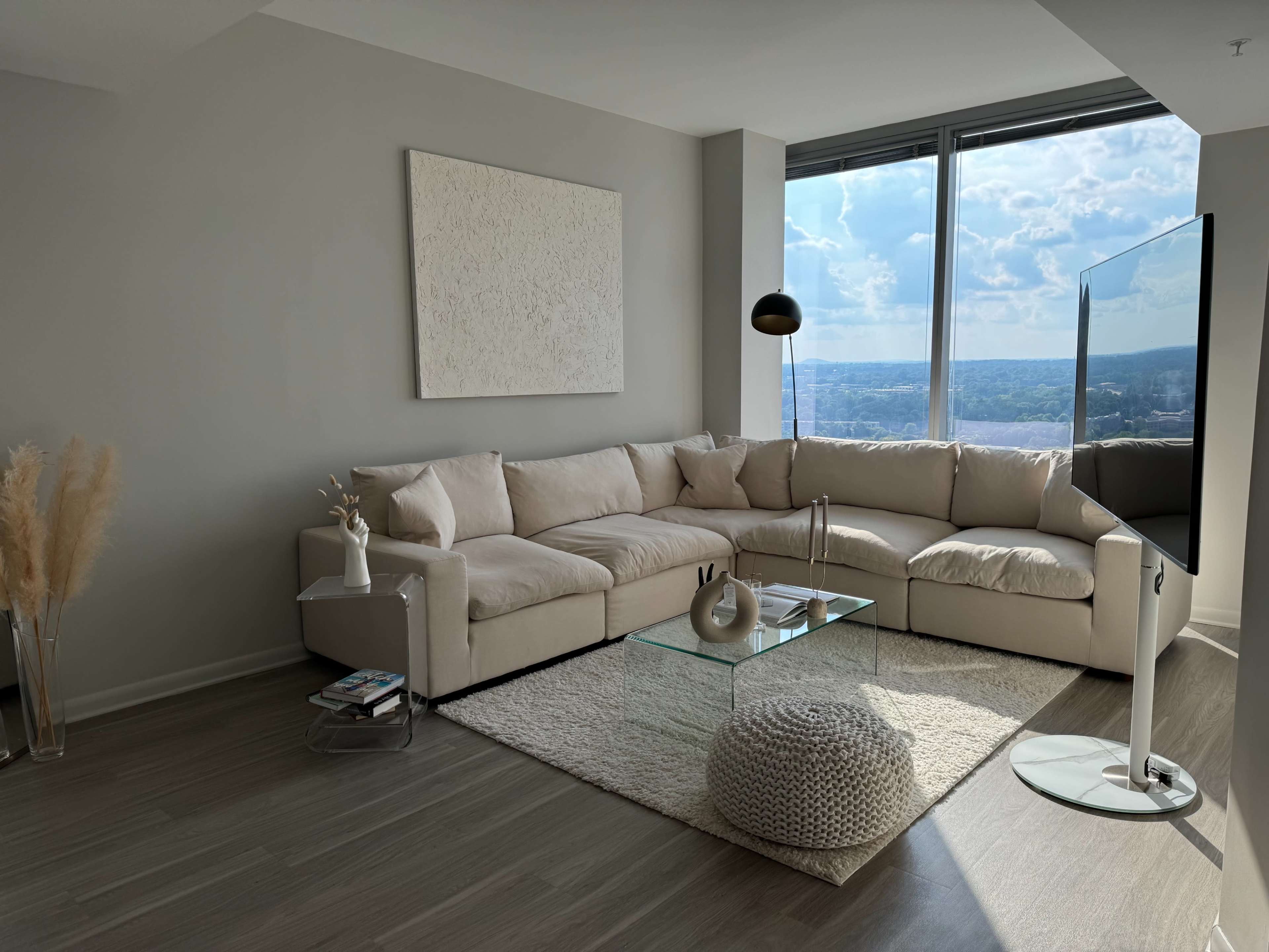 A light-filled living room featuring a large beige sectional sofa, a glass coffee table, and a flat-screen TV positioned near a window with a view.