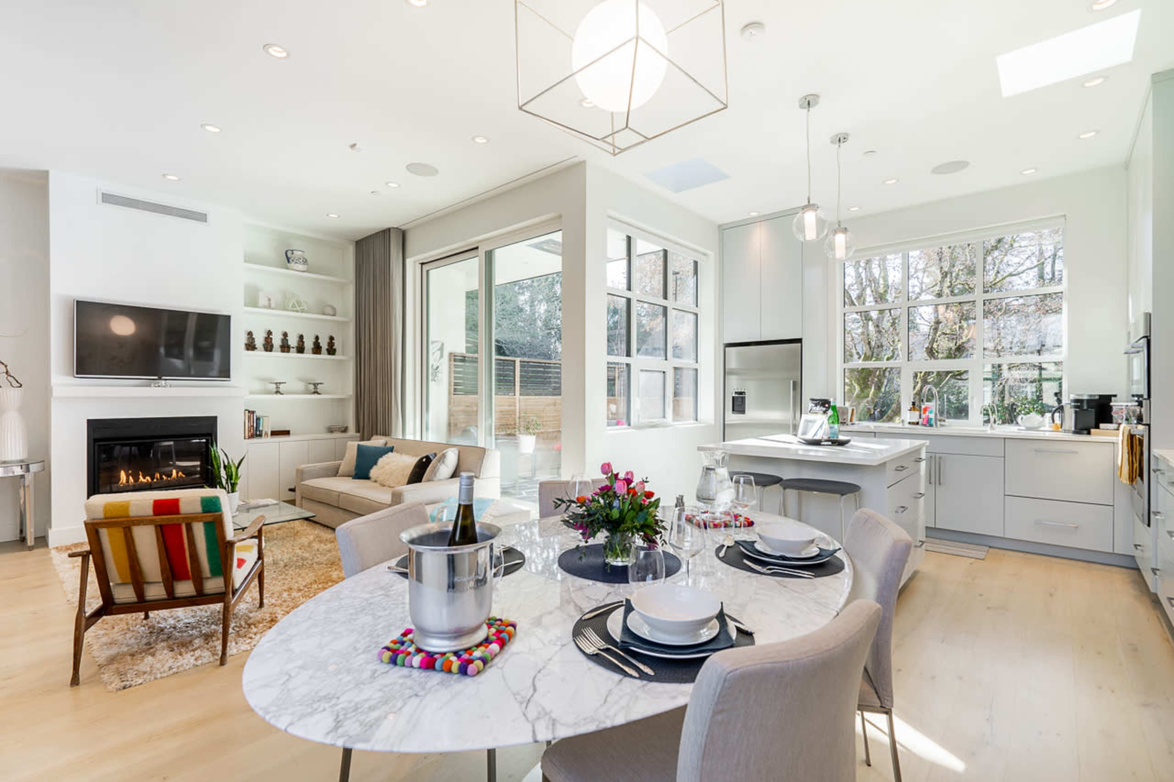 The image shows a modern kitchen and living area featuring a round marble dining table, a cozy sofa, and large windows with a view to an outdoor space.