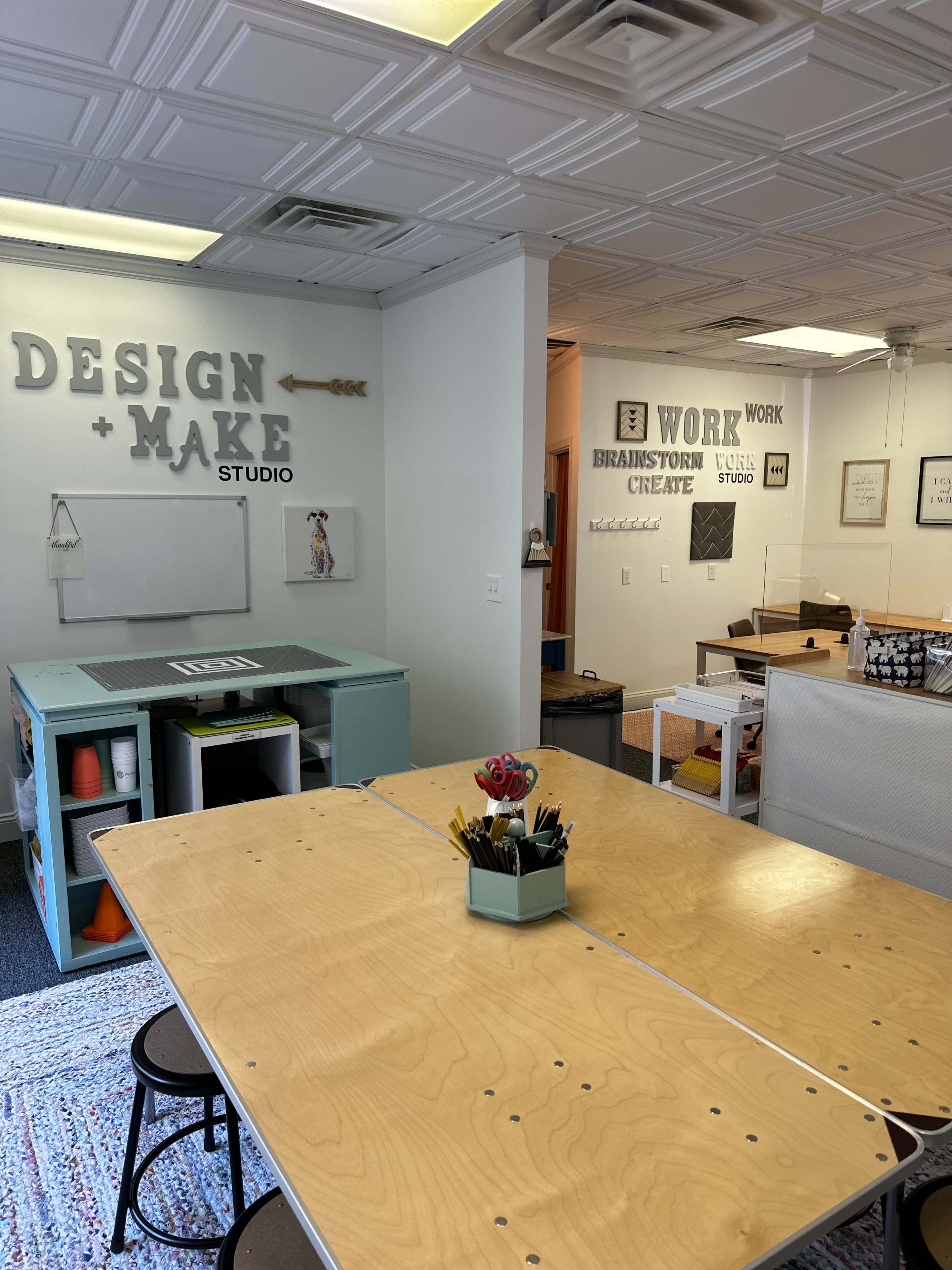 A creative workspace with a large wooden table at the center, surrounded by a colorful storage unit and various work areas featuring inspirational signage.