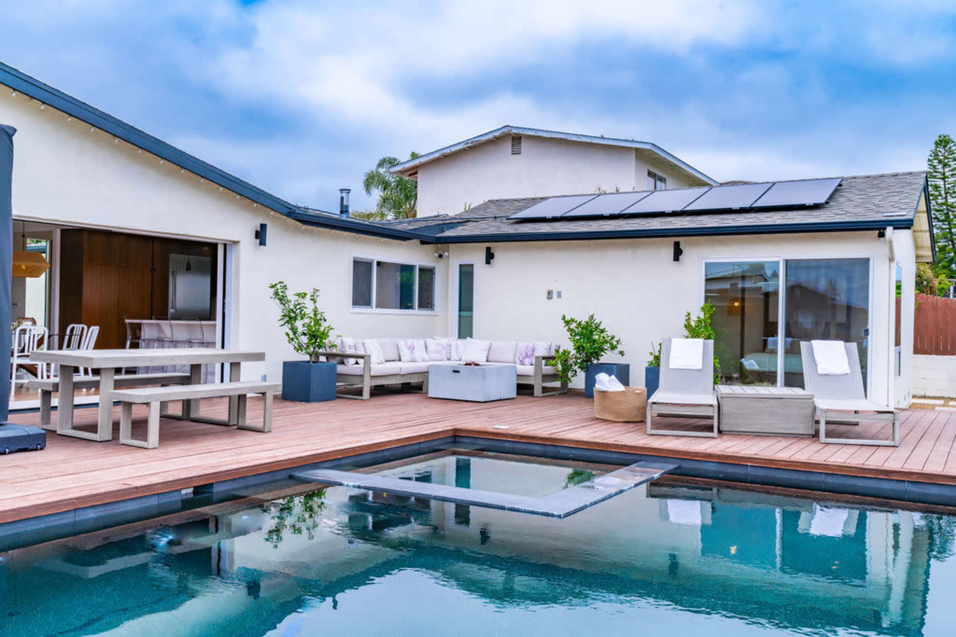 The image shows a modern backyard with a swimming pool, wooden deck, lounge chairs, and a dining table adjacent to a house equipped with solar panels.