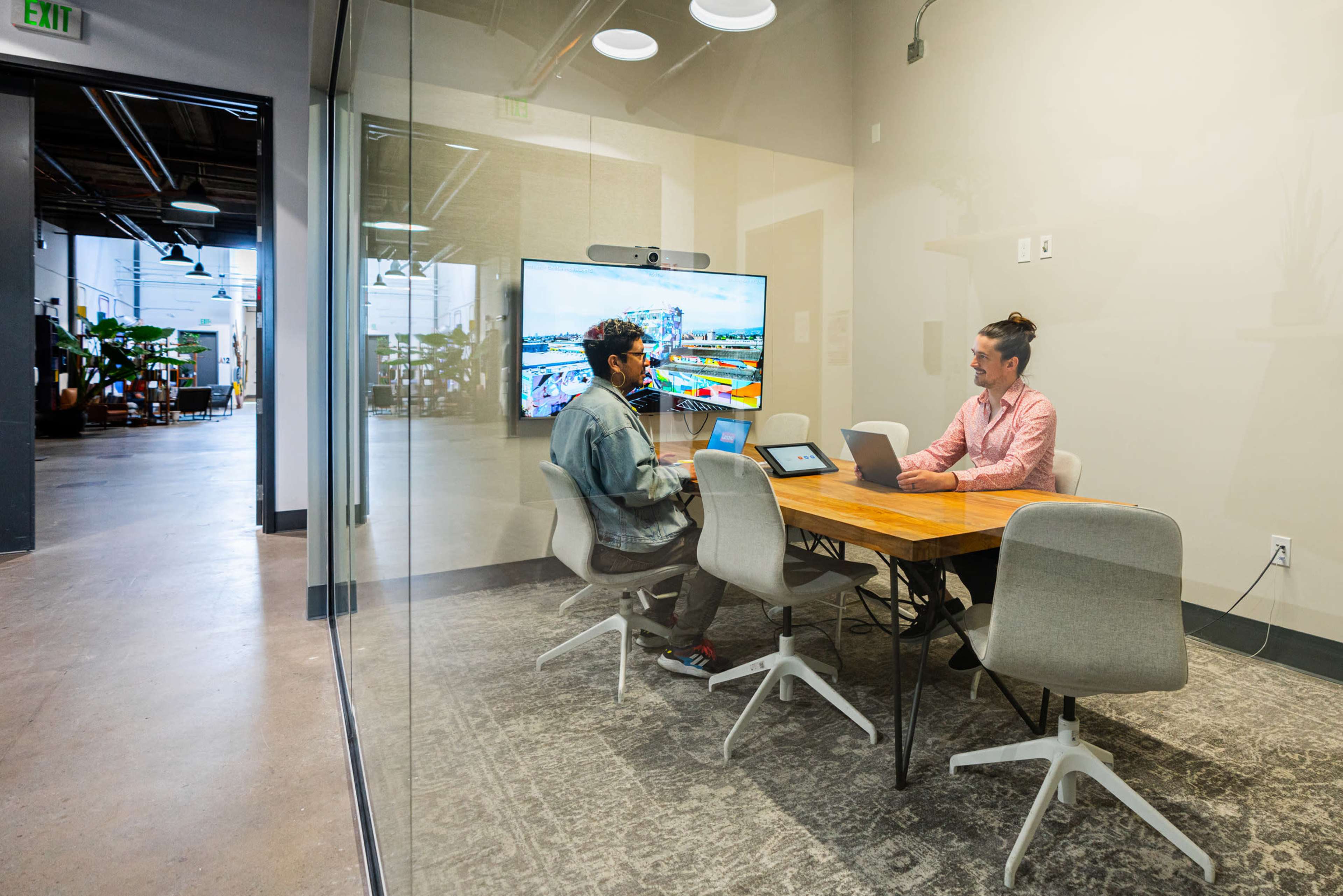 Two people sit at a table in a glass-walled conference room, working on laptops with a large screen displaying a colorful presentation in the background.