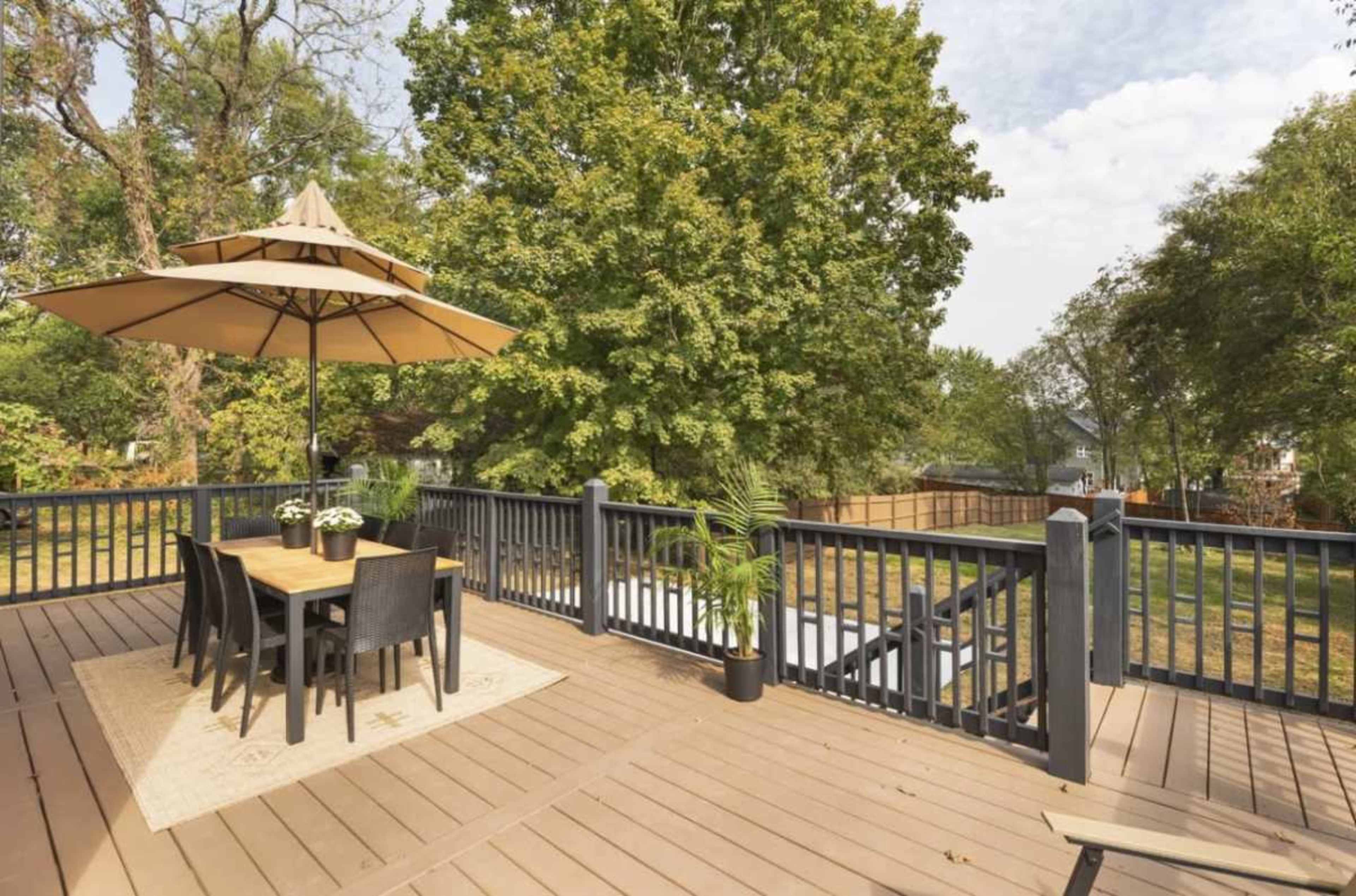 A wooden deck features a dining table with chairs under a large umbrella, surrounded by trees and a grassy area.
