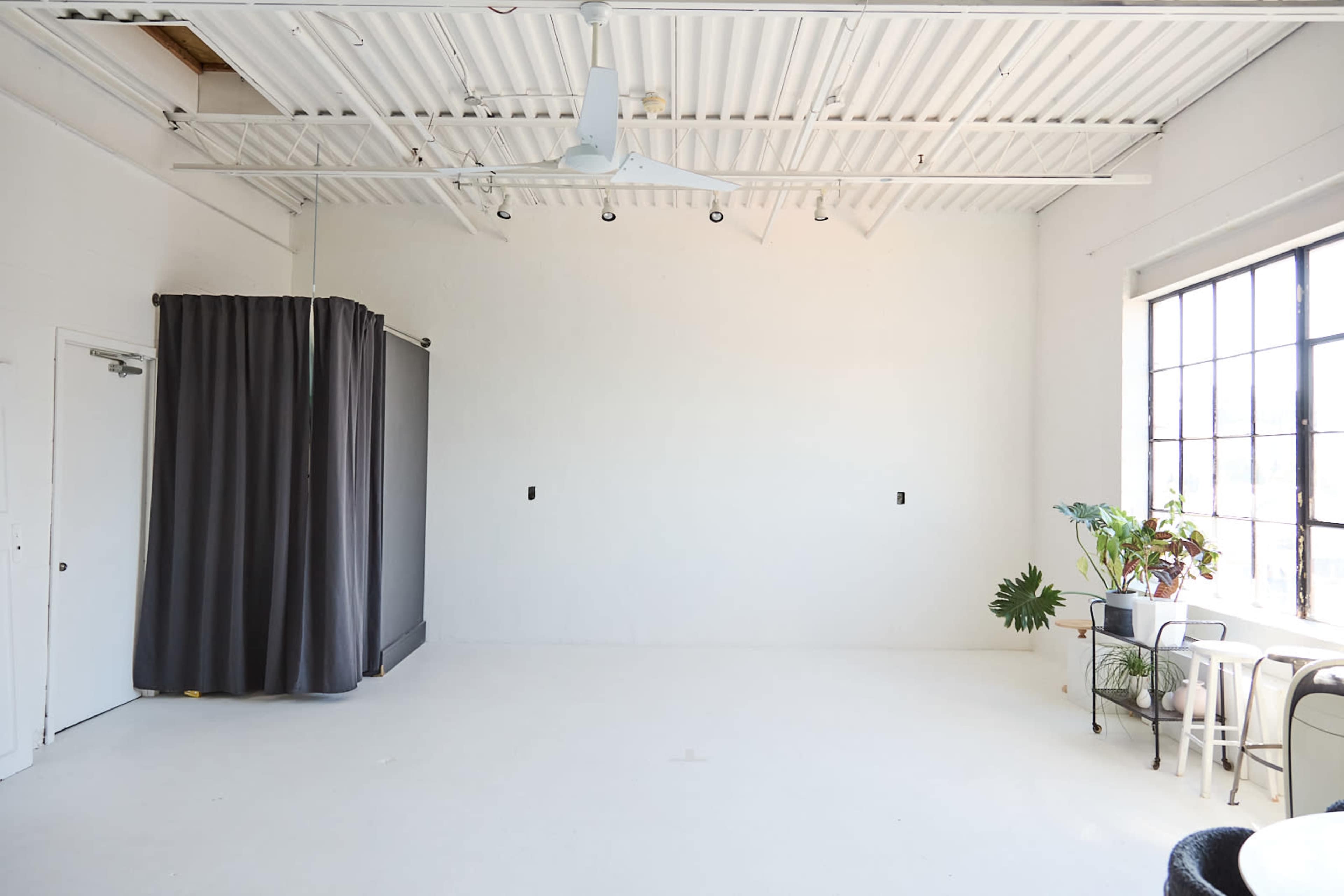 The image shows a minimalist, empty room with a gray curtain on one side, a window allowing natural light, and a small table with chairs and plants near the wall.