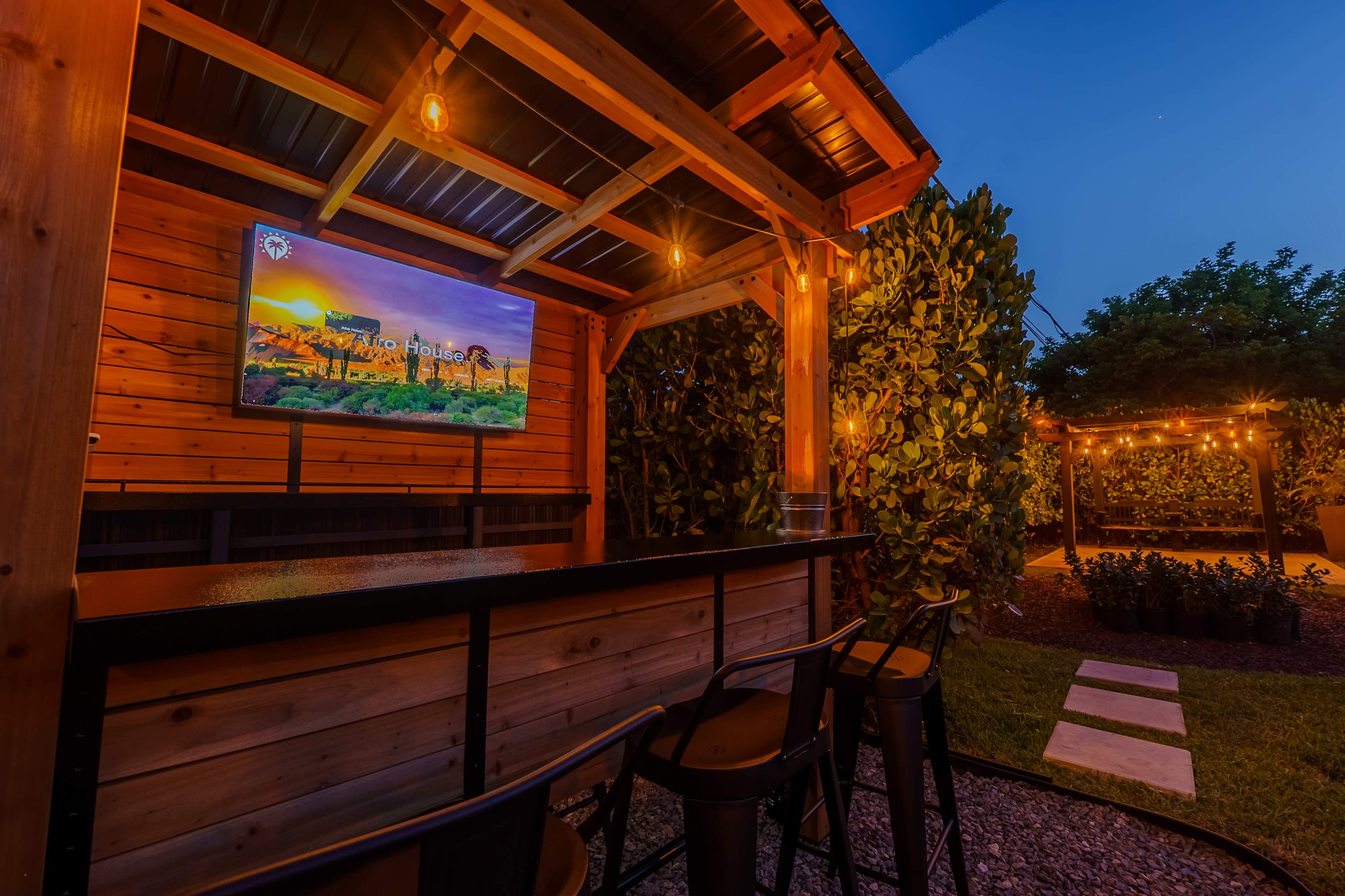 A wooden outdoor bar area features a television mounted on the wall, surrounded by string lights and a garden in the background.