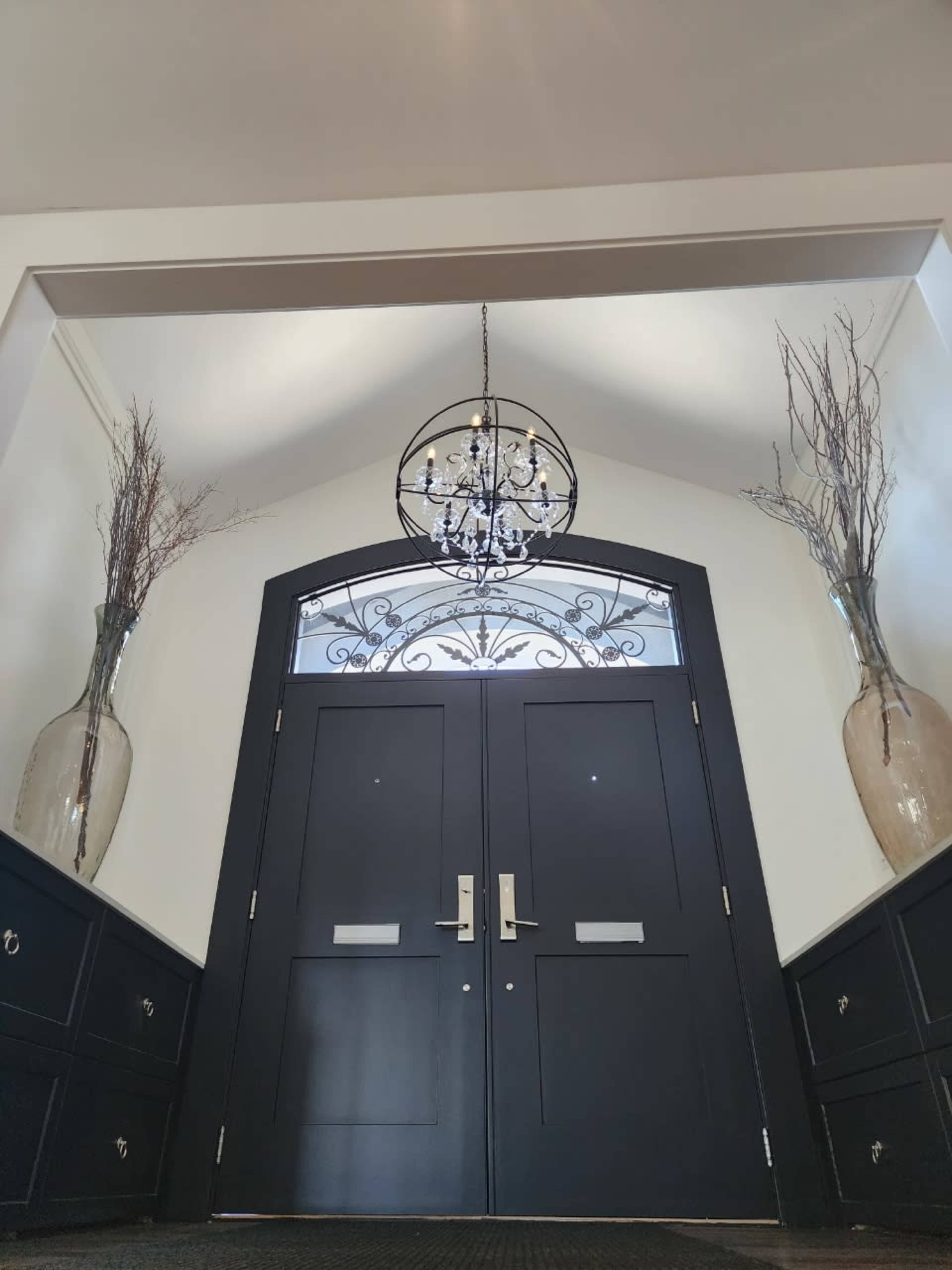 A spacious entryway featuring a pair of double black doors, a decorative chandelier overhead, and tall vases with branches on each side.