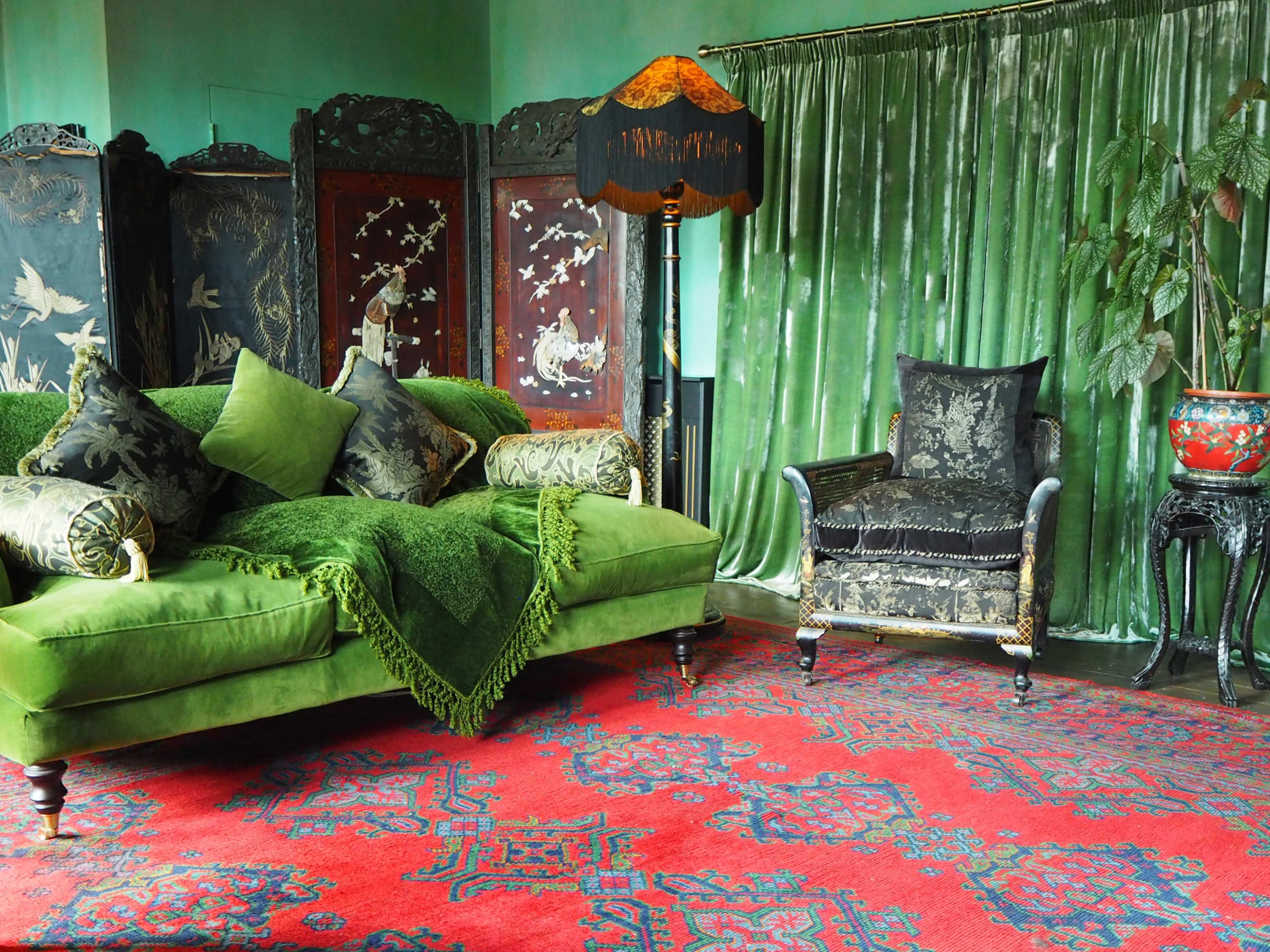 A green sofa adorned with decorative pillows, a matching armchair, and a dark red patterned carpet, set against a backdrop of green curtains and ornate room dividers.