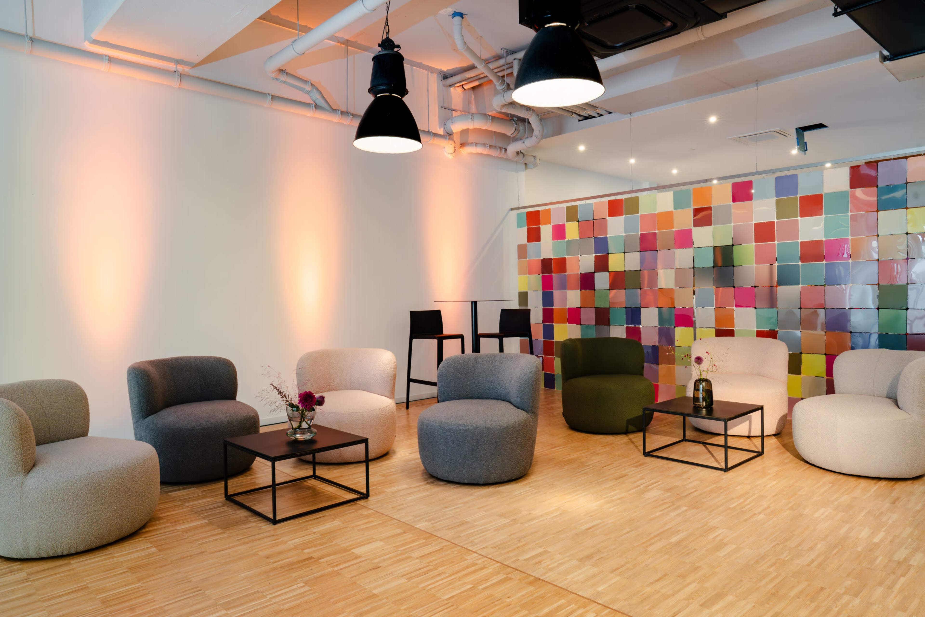 The image shows a modern lounge area with several upholstered chairs arranged around black tables, accompanied by colorful tiled wall decor and soft lighting.