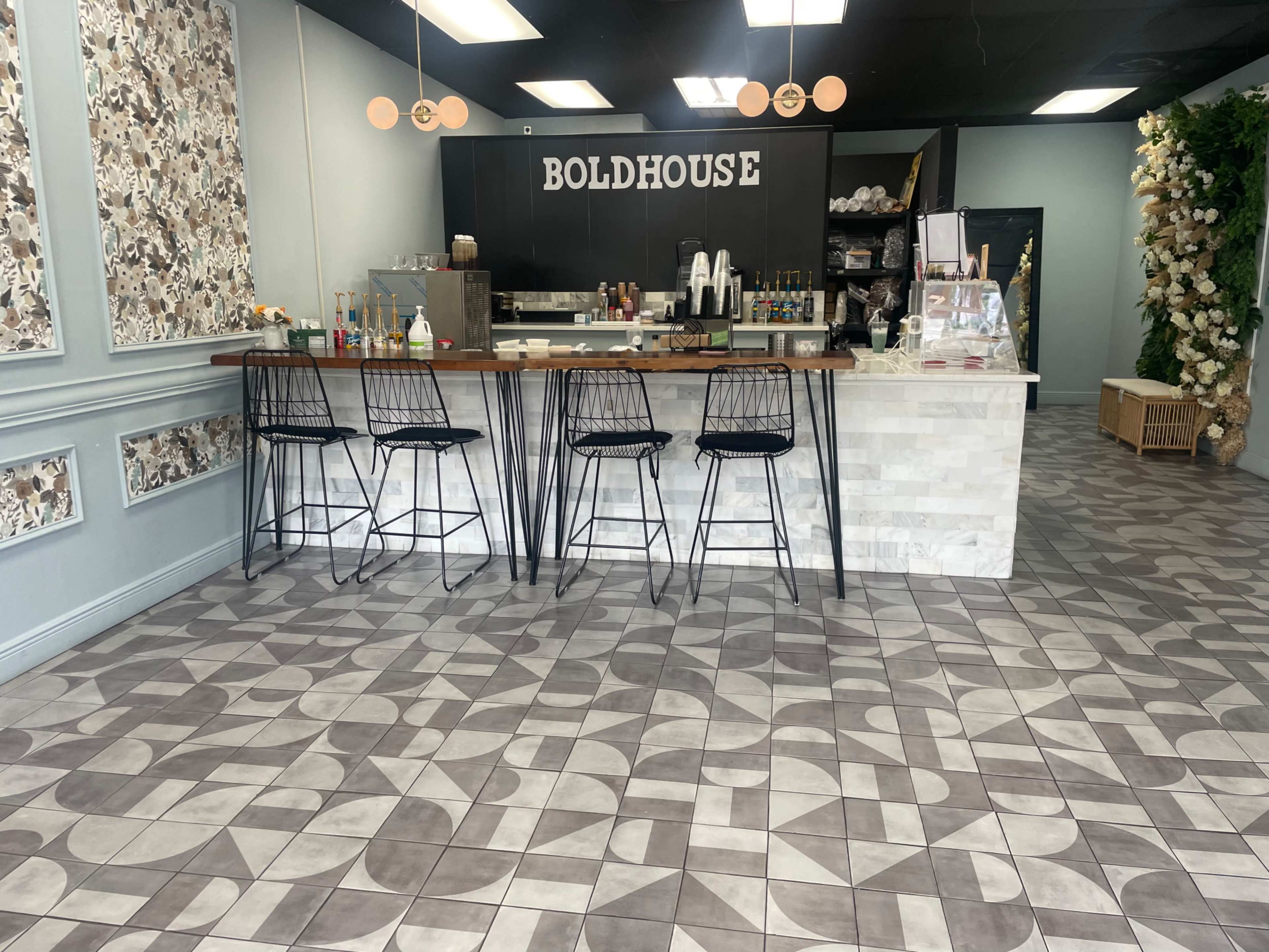 The image shows a café space with a modern counter, four black wire bar stools, and patterned flooring, decorated with artwork and floral elements.