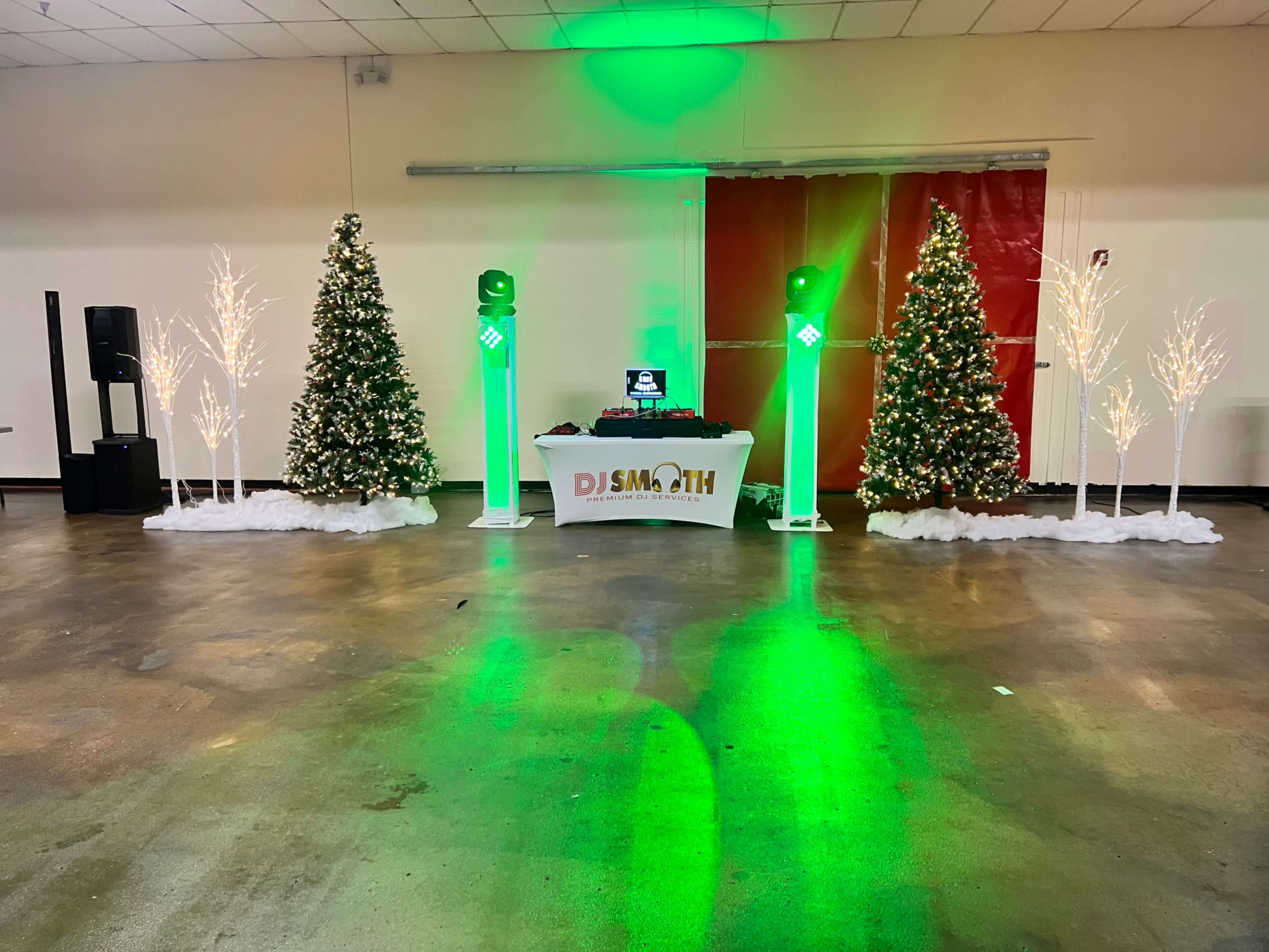 A DJ setup is displayed in a decorated space with two Christmas trees, illuminated columns, and artificial snow on the ground.