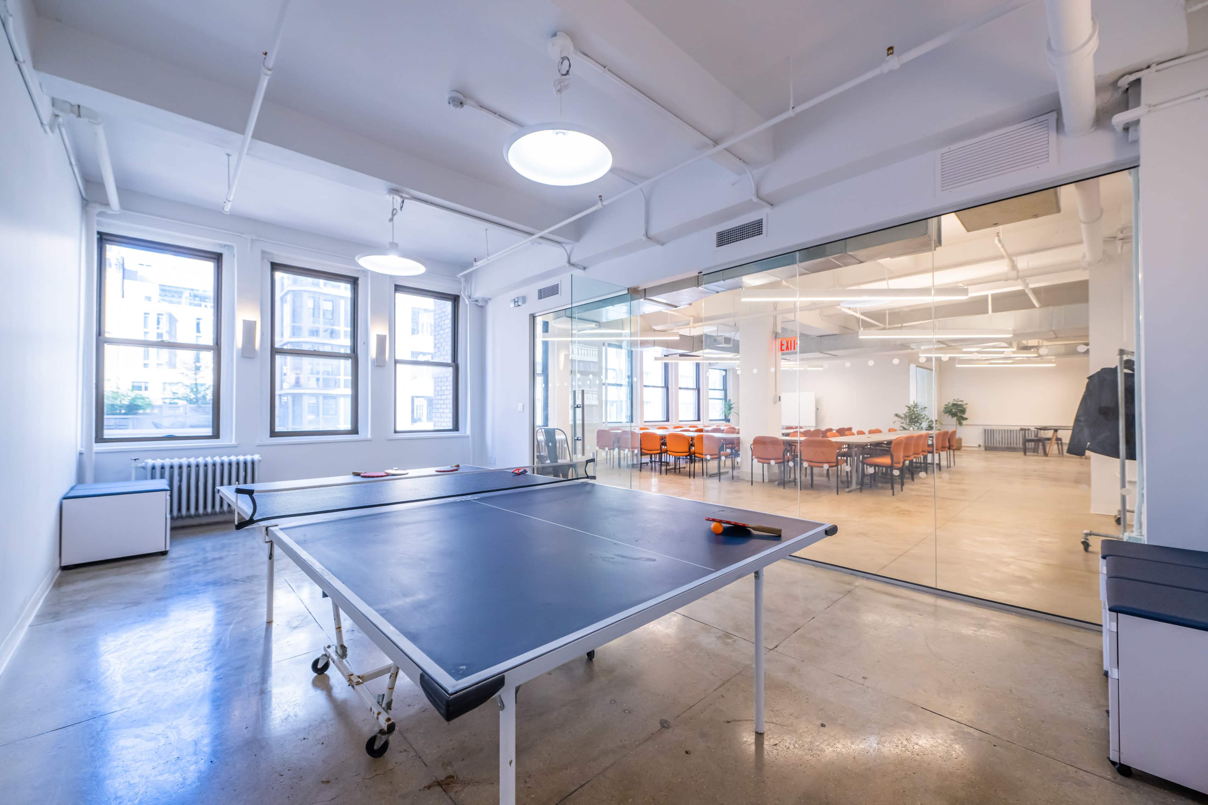 An open office space featuring a ping pong table in the foreground and a conference area with a long table and chairs visible through glass walls in the background.
