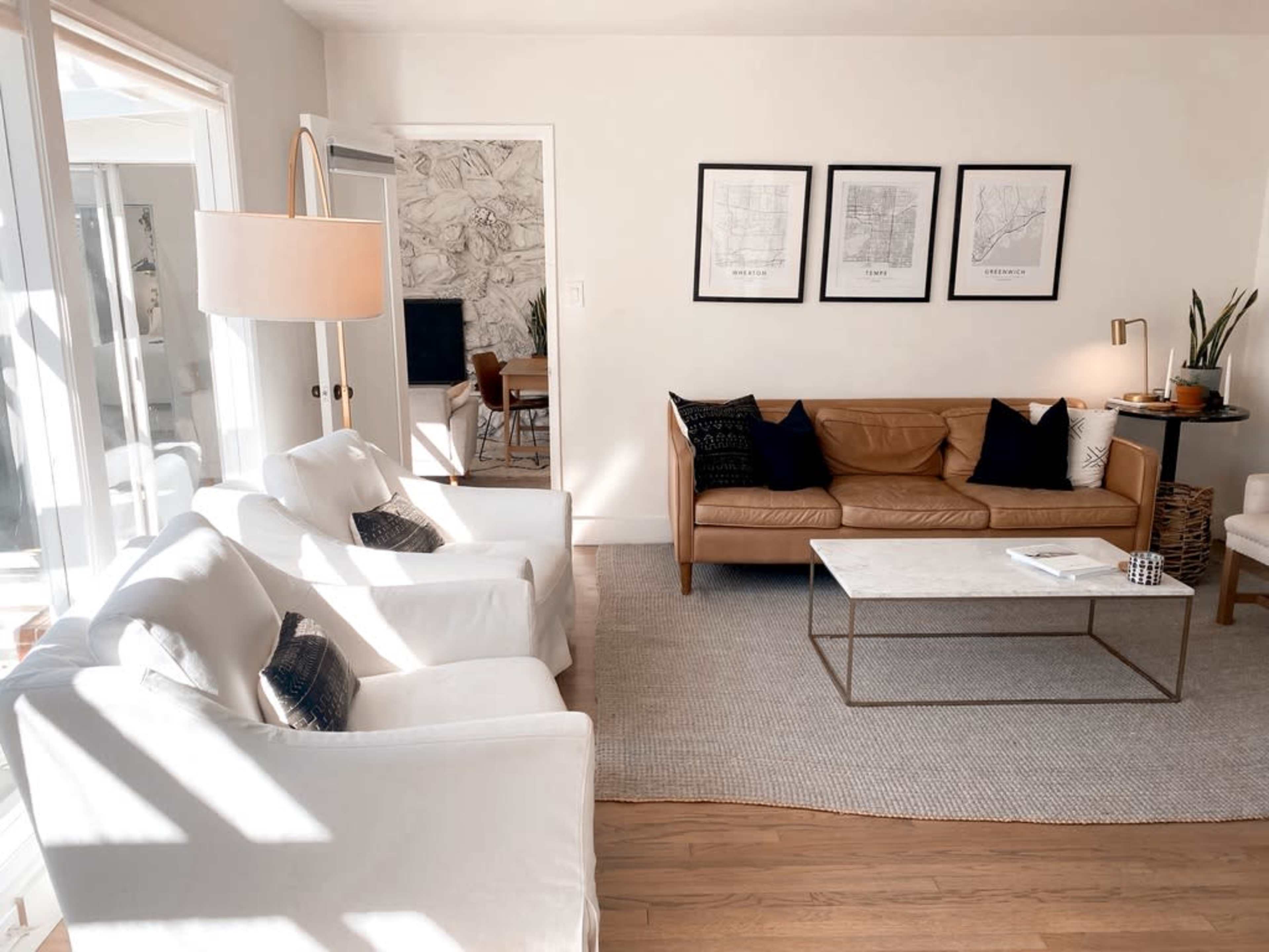 A bright living room features a leather sofa, two white armchairs, a marble coffee table, and framed artwork on the walls.