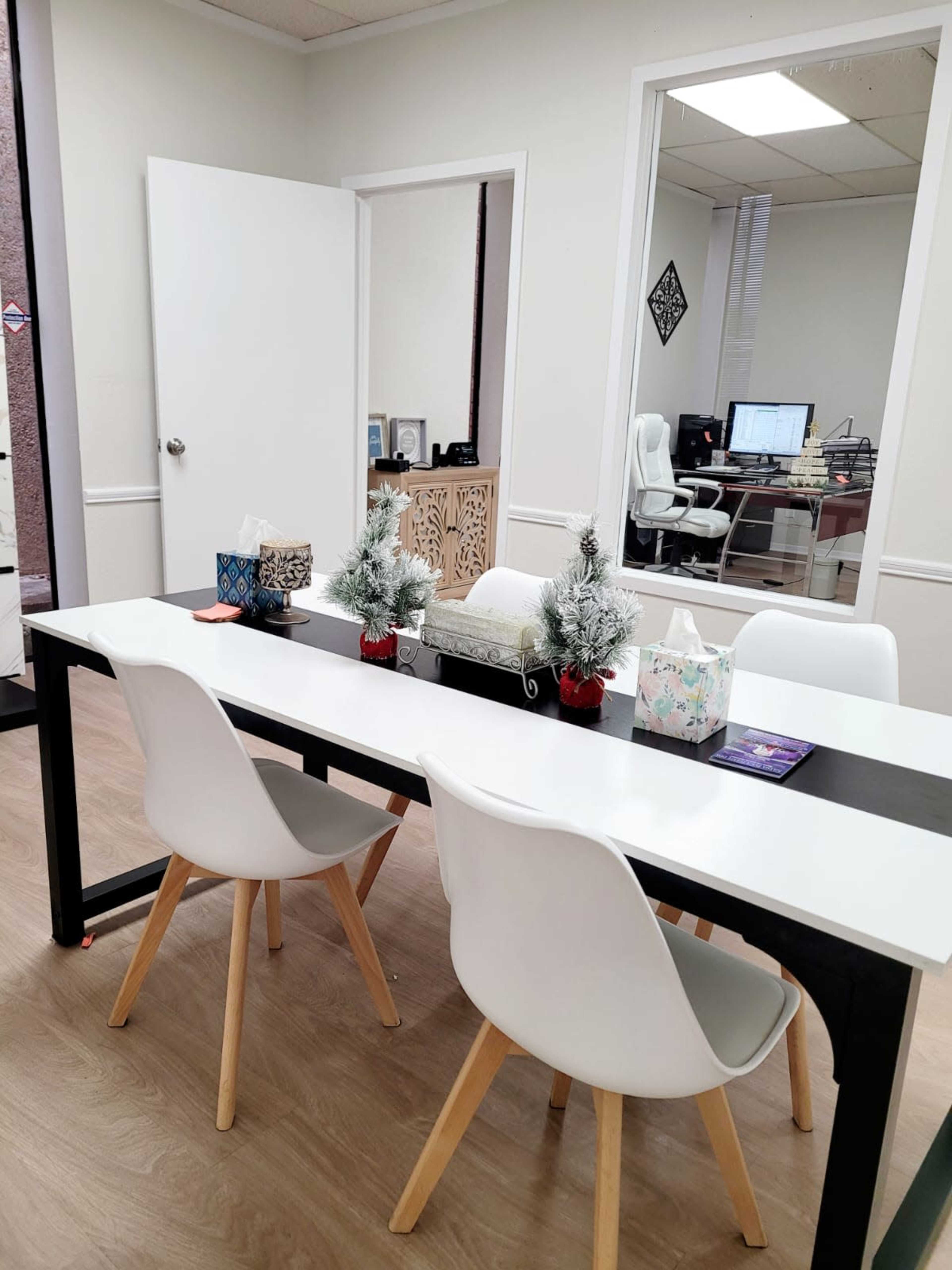 A modern office space features a rectangular table surrounded by four white chairs, adorned with small evergreen decorations and tissue boxes, with a separate workspace visible through a glass window.