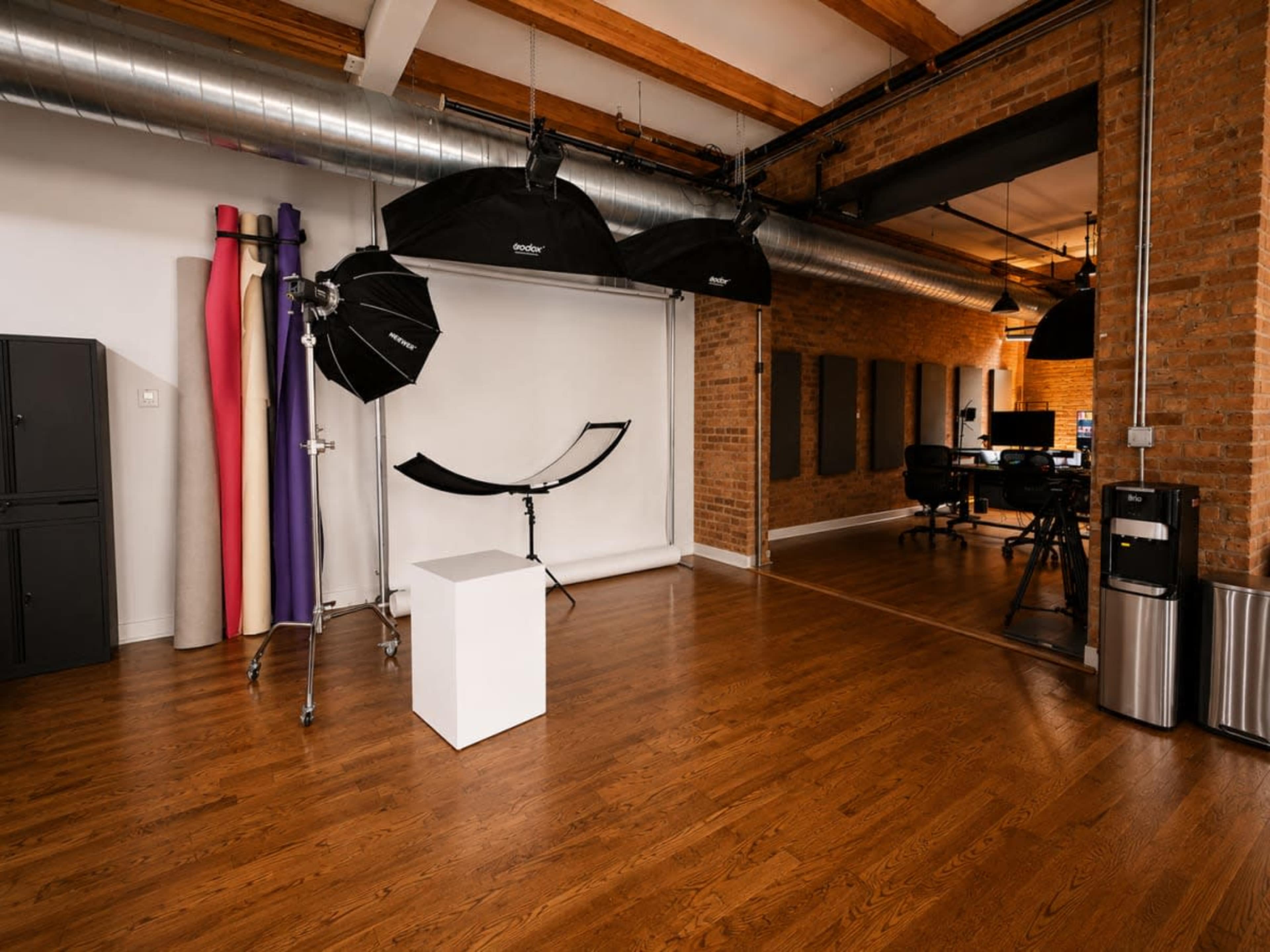 The image shows a photography studio with a white background, lighting equipment, a small white pedestal, and colorful backdrops on one side.