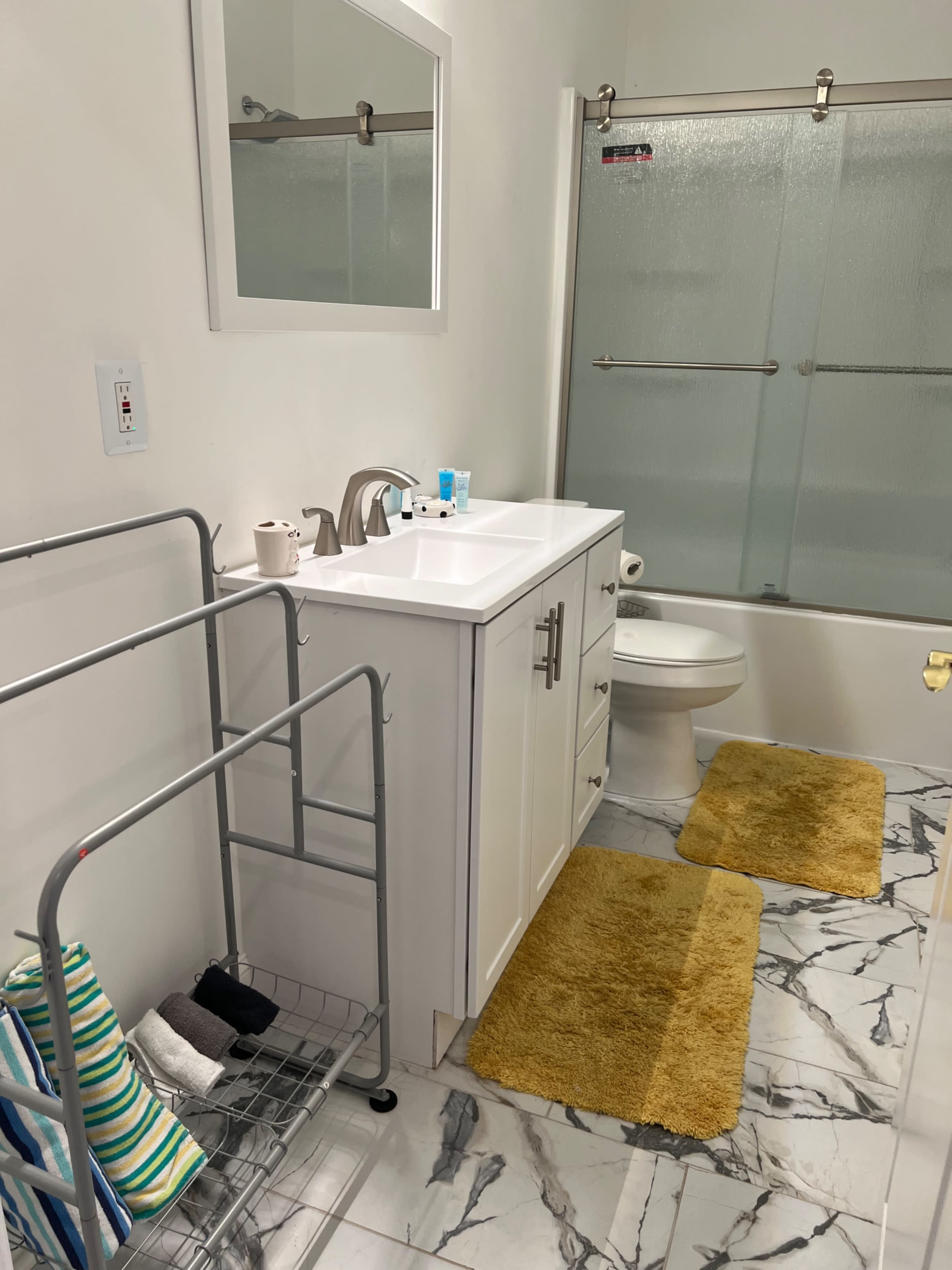 A clean bathroom features a white vanity with a sink, a toilet, and a shower with sliding glass doors, alongside a towel rack and yellow bath mats on the floor.