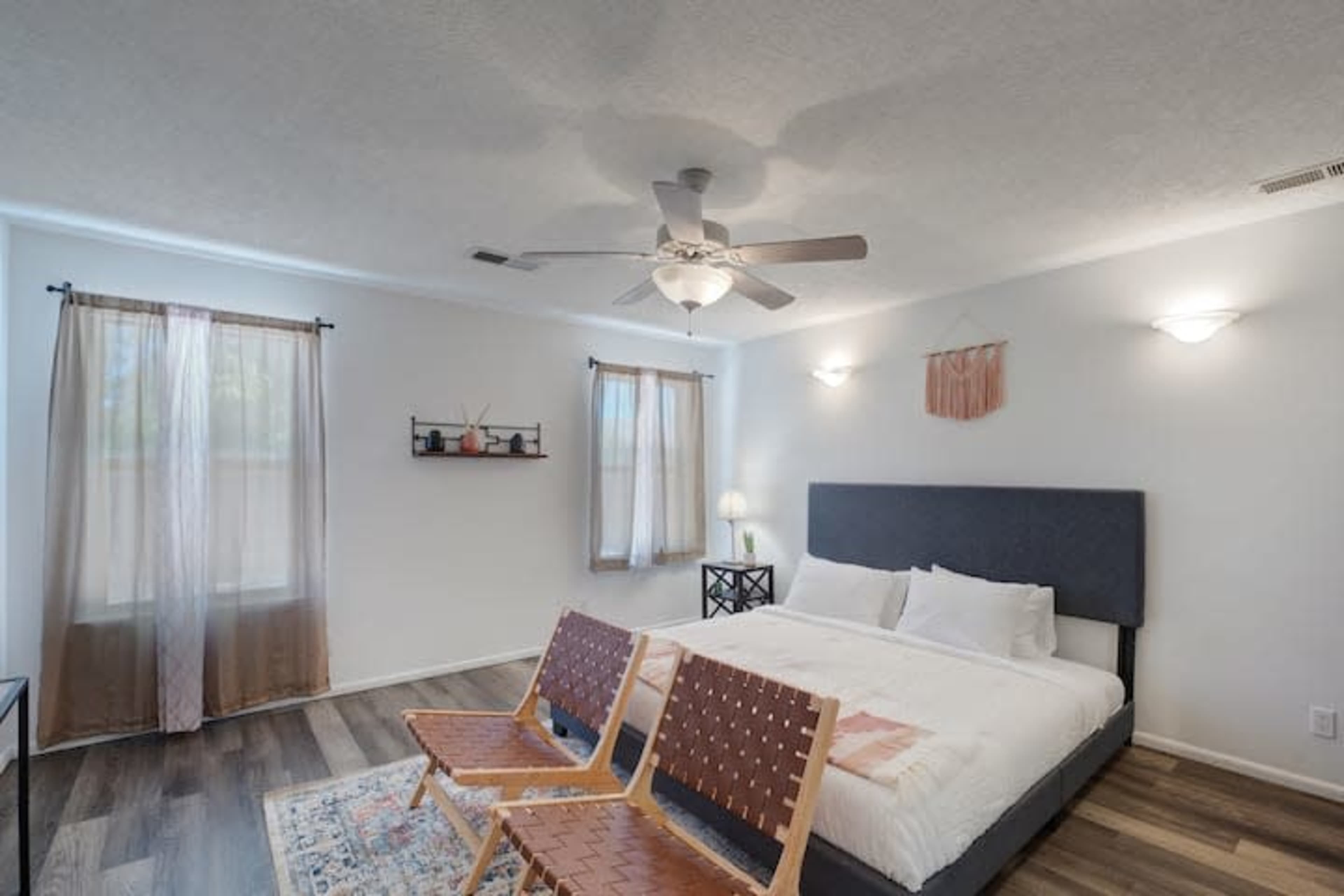 A neutral-toned bedroom with a large bed, two wooden chairs, and light curtains allowing natural light to enter.