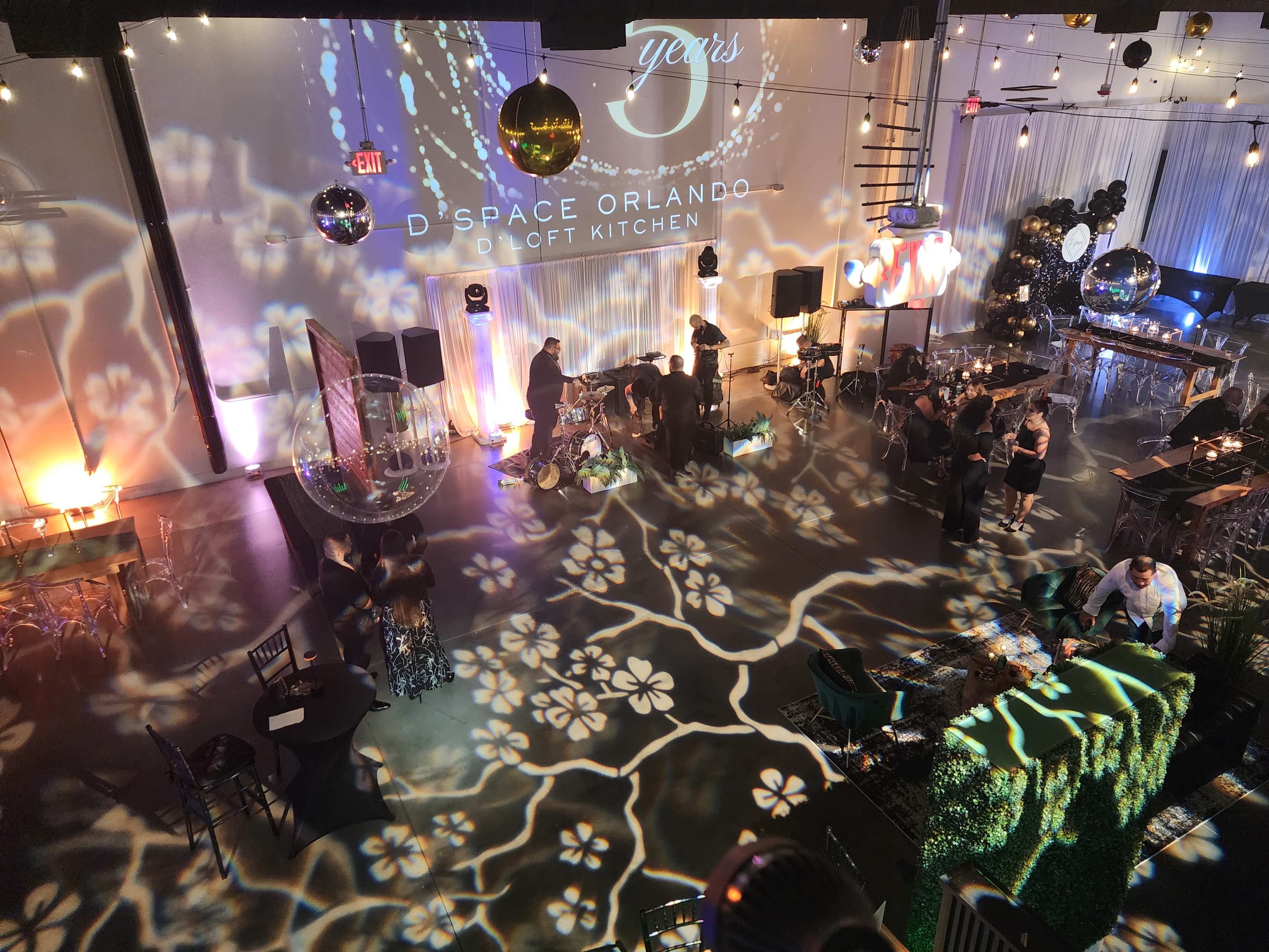 The image shows a partially decorated event space with guests mingling, surrounded by colorful lighting and floral patterns projected on the walls and floor.