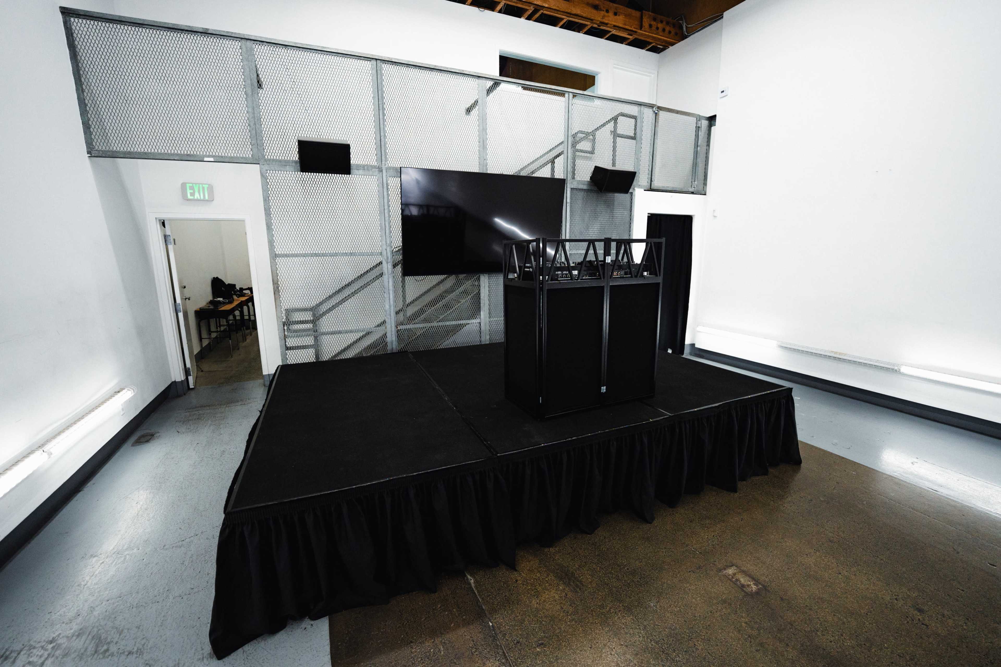 The image shows a stage setup with a black podium and a large screen in a sparse, well-lit room.