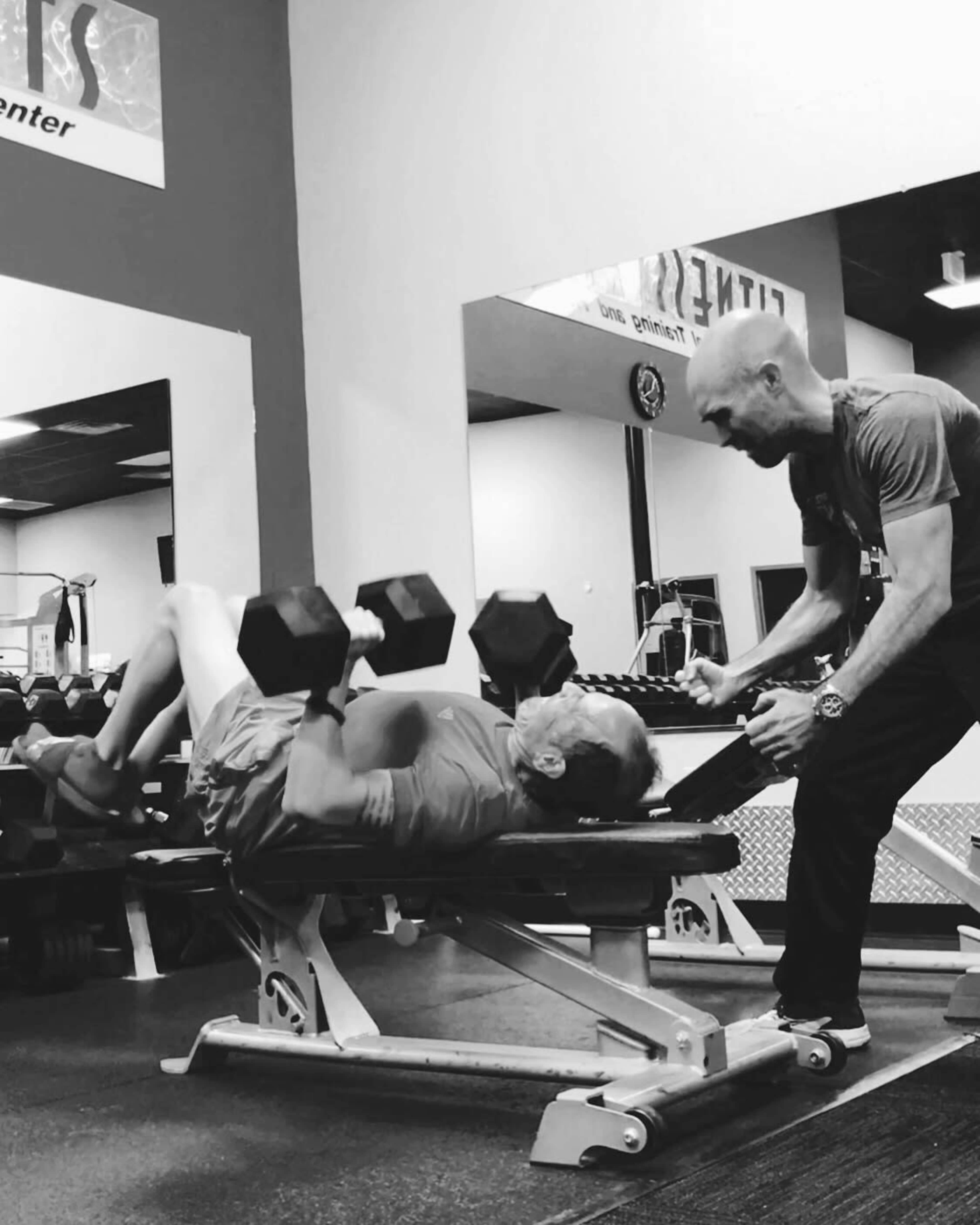 A trainer assists a man lifting dumbbells while lying on a bench in a gym.