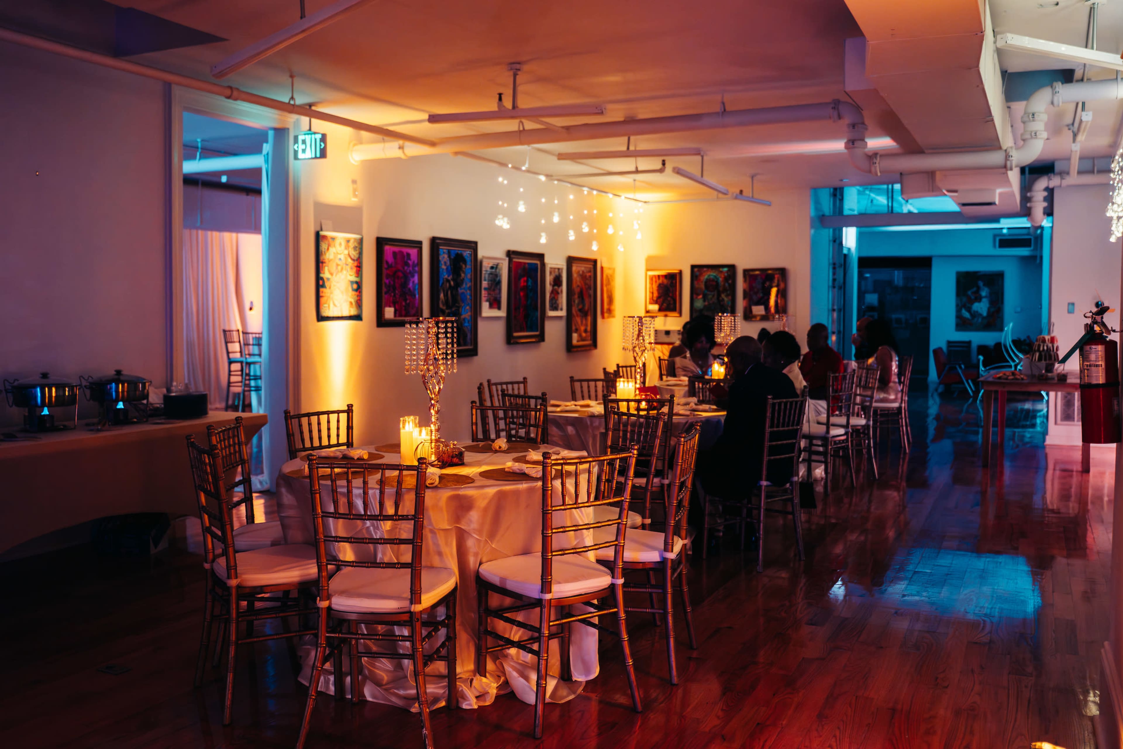 A dining area with round tables and chairs is illuminated by soft lighting, featuring artwork on the walls and several guests seated at the tables.