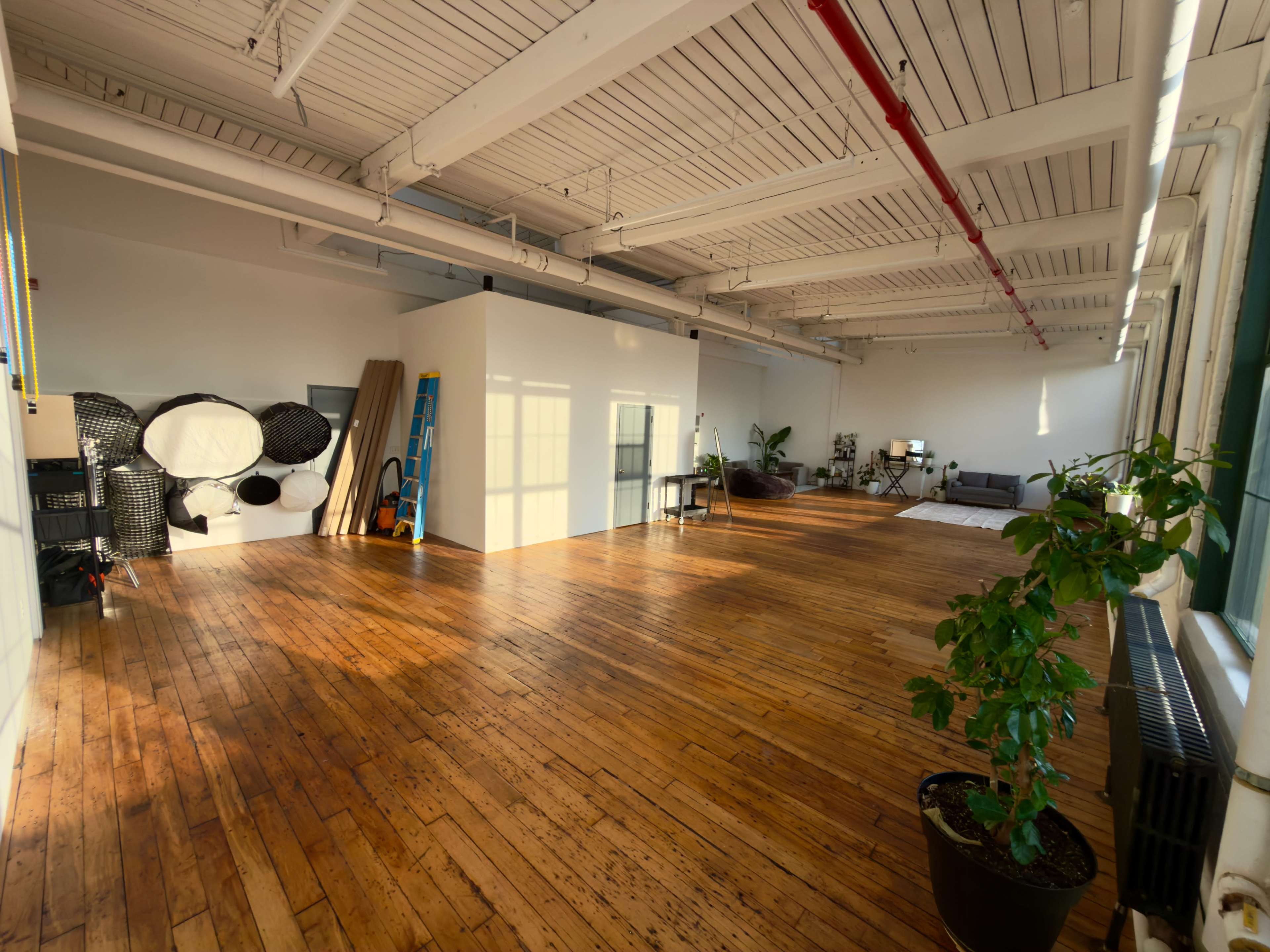 The image shows a spacious, sunlit studio with wooden flooring, a few pieces of furniture, and a ladder against a wall.