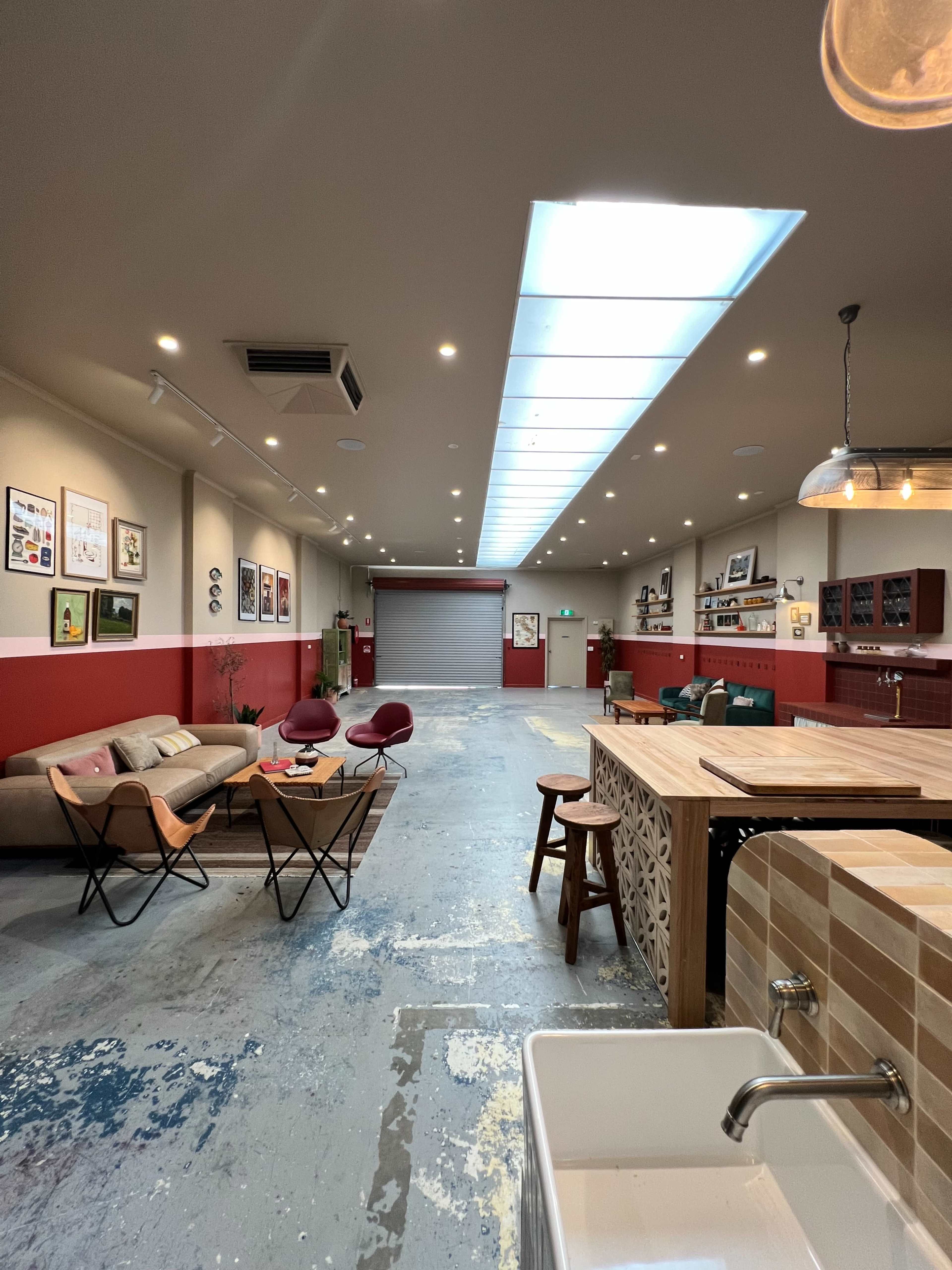 The image shows a spacious room with a long skylight, featuring a lounge area with couches, chairs, and a wooden table, along with a kitchen area and a garage door at one end.