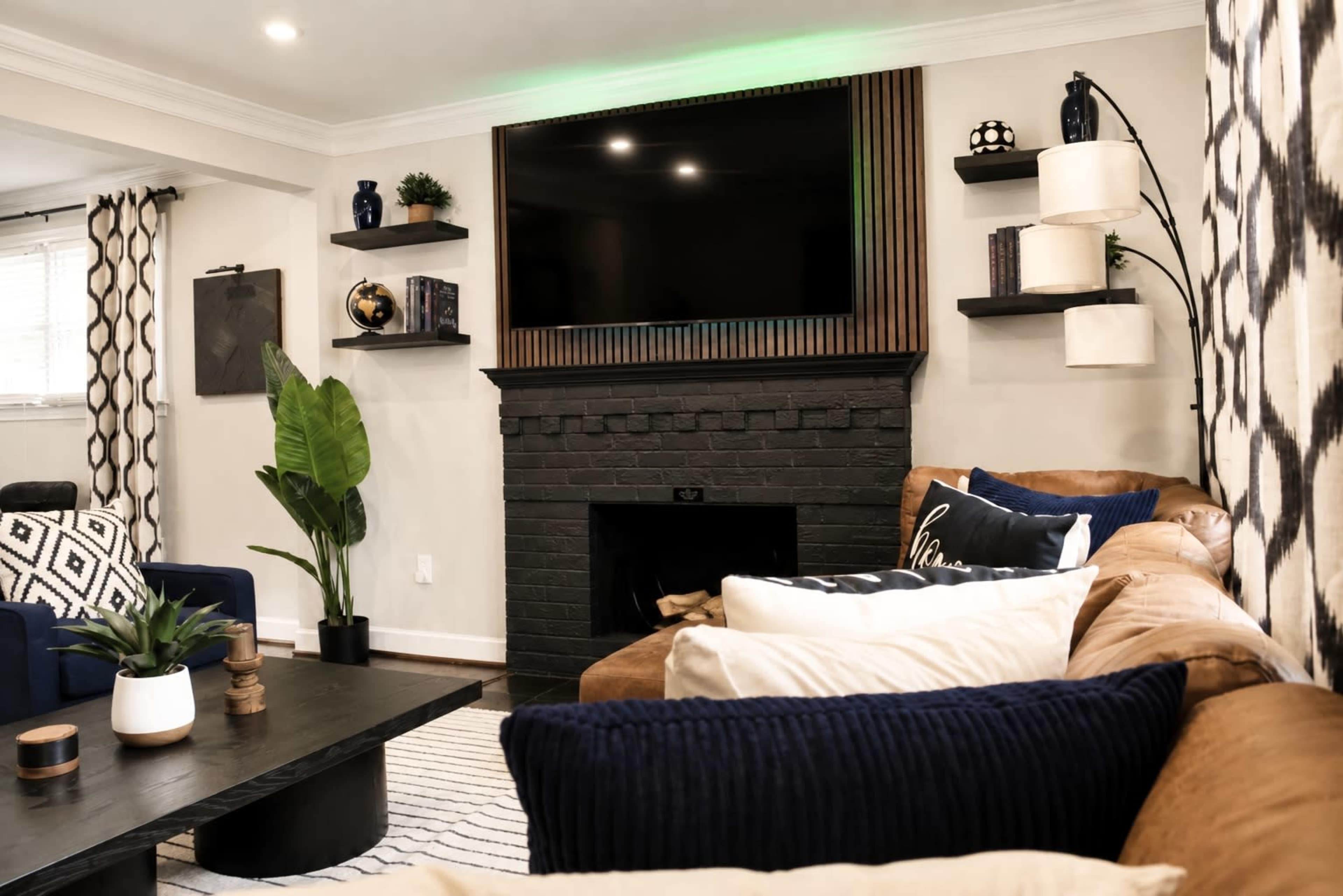 The image shows a living room featuring a dark brick fireplace, a mounted flat-screen TV above it, and a mix of modern furniture, including a blue couch and a brown leather sectional.