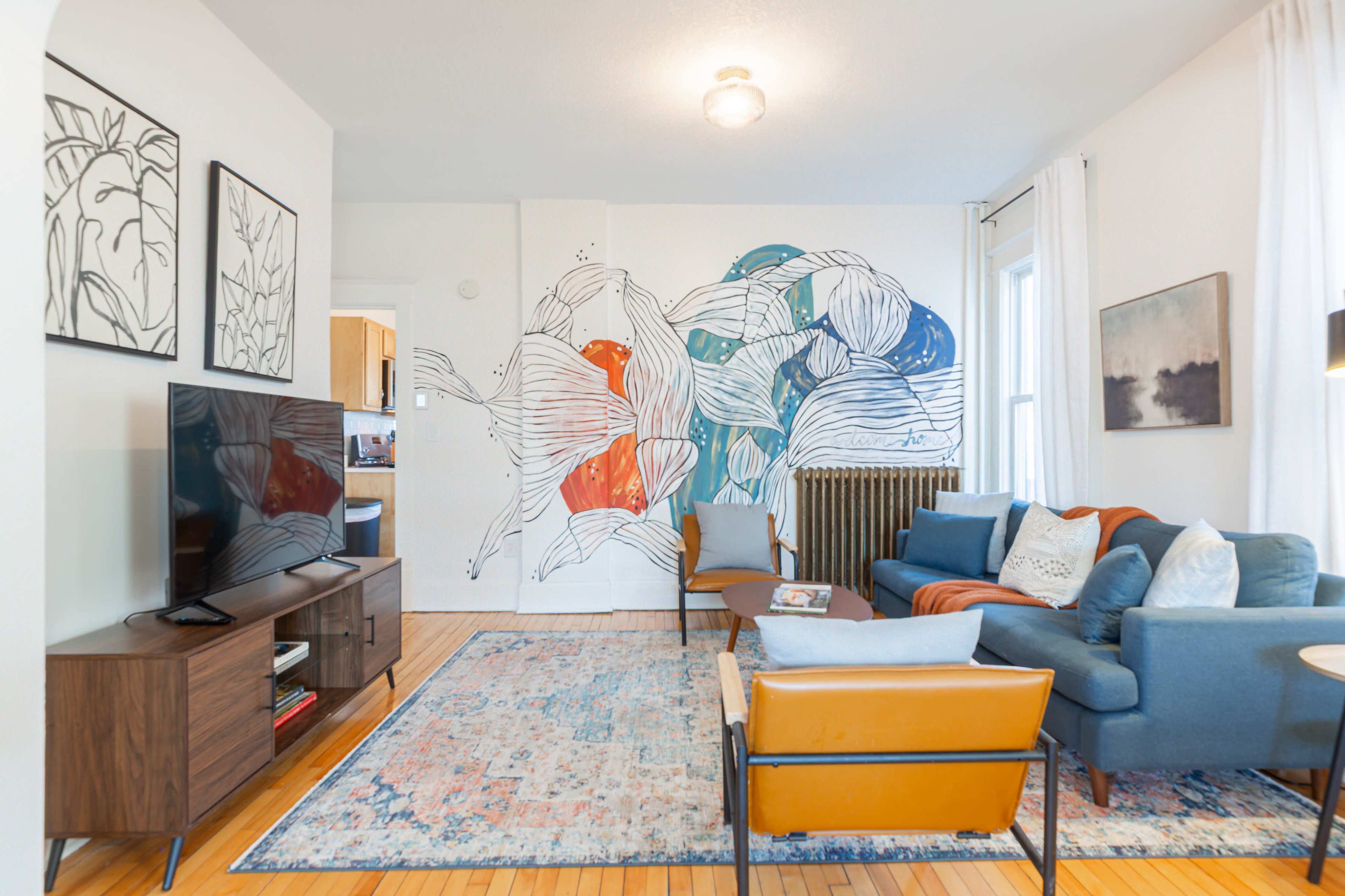 A living room features a blue couch, an orange chair, a wooden media console with a television, a decorative area rug, and a mural of abstract flowers on the wall.