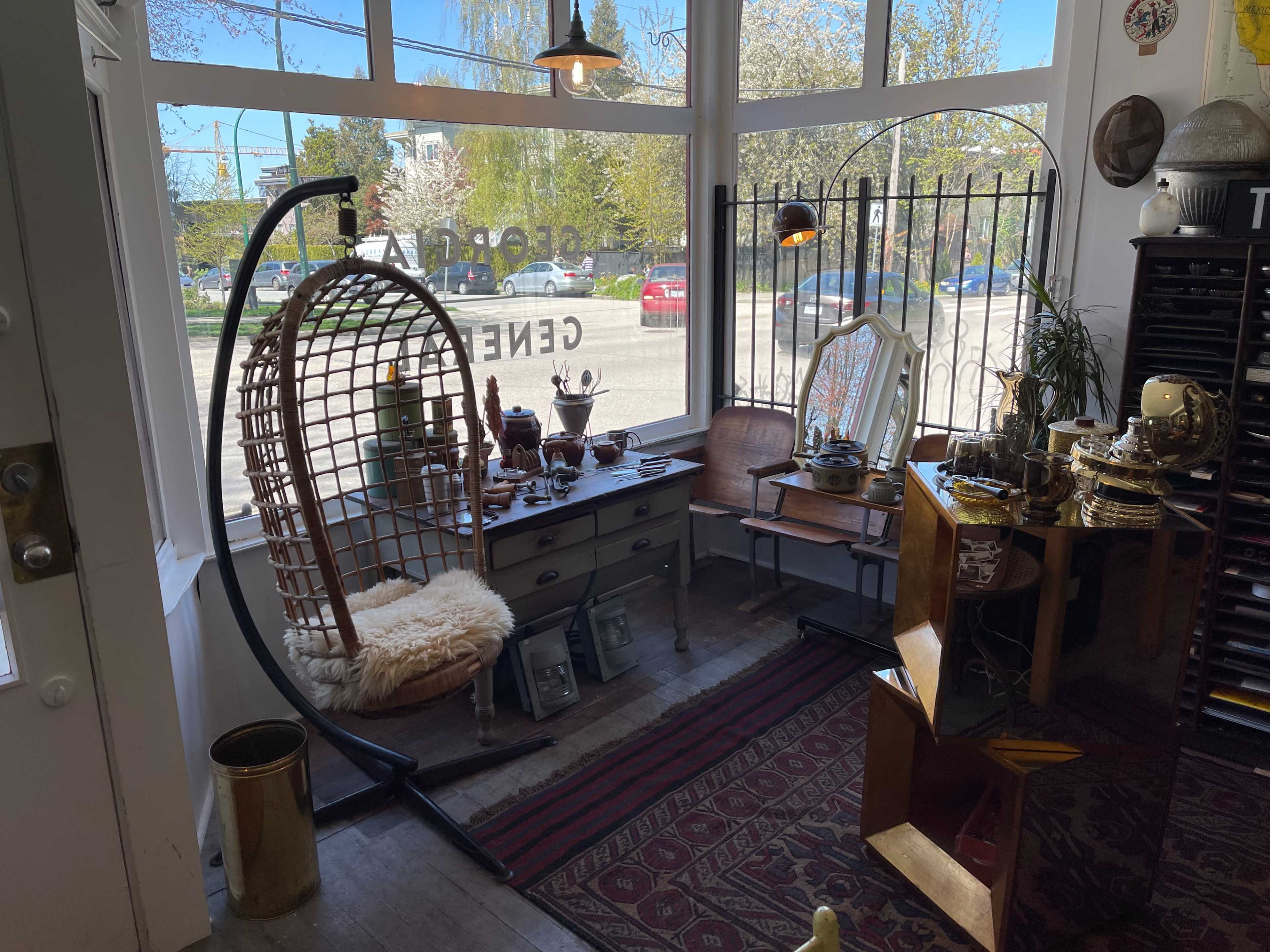 A brightly lit vintage shop interior featuring a hanging wicker chair, a table with various decorative items, and shelves filled with antiques.