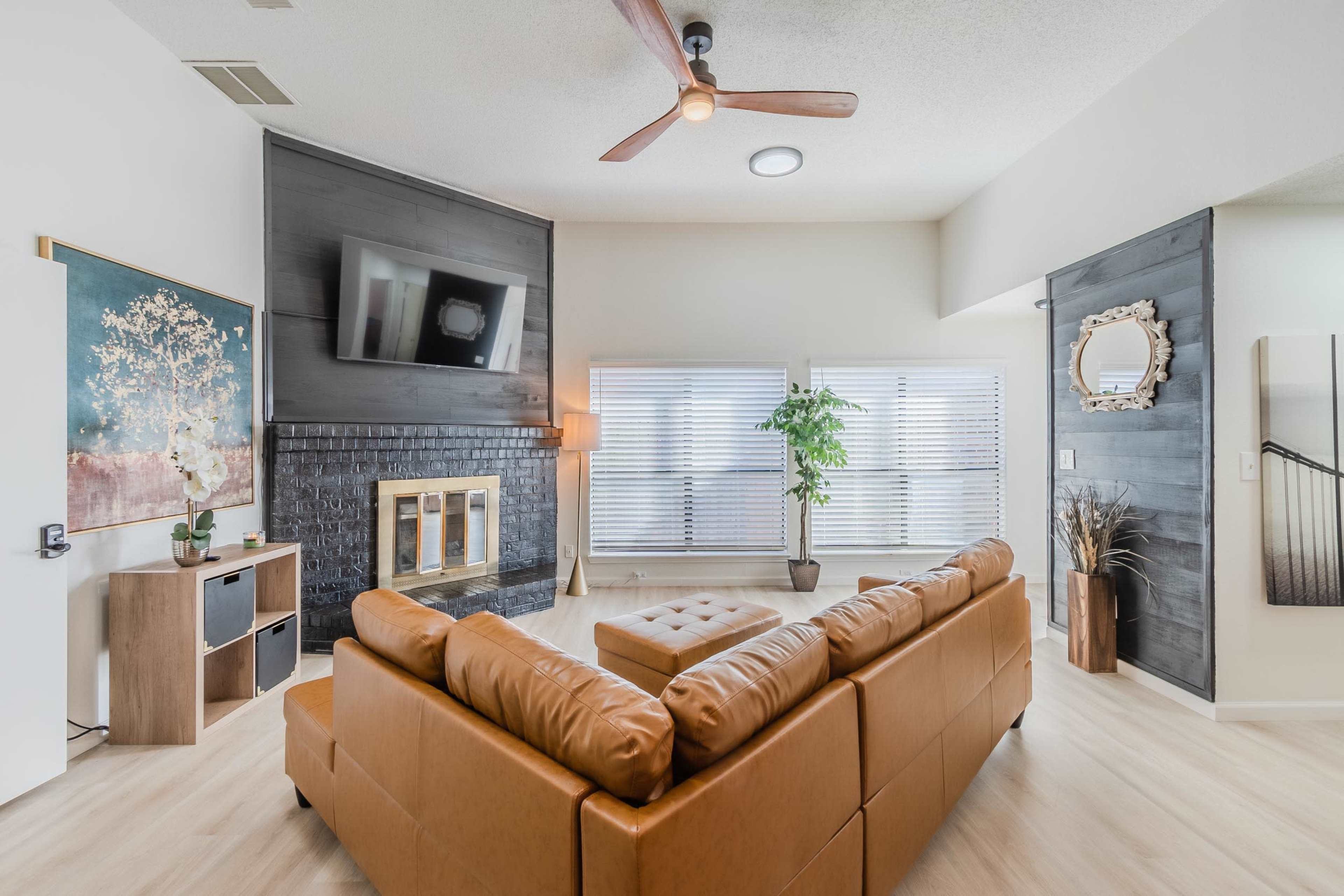 A modern living room featuring a brown leather sectional sofa, a wall-mounted television, and large windows with blinds.