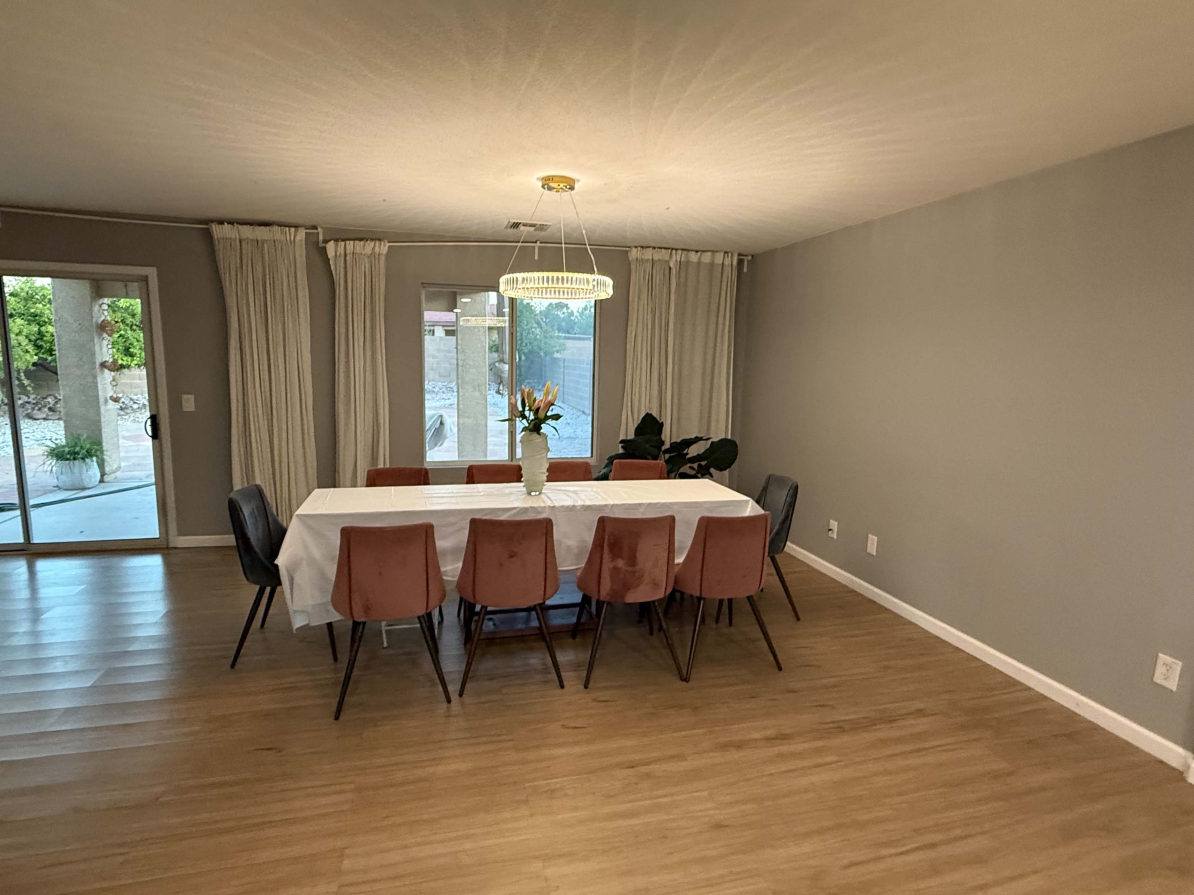 A large dining table is set with a white tablecloth and surrounded by eight chairs in a brightly lit room with large windows and sliding doors.
