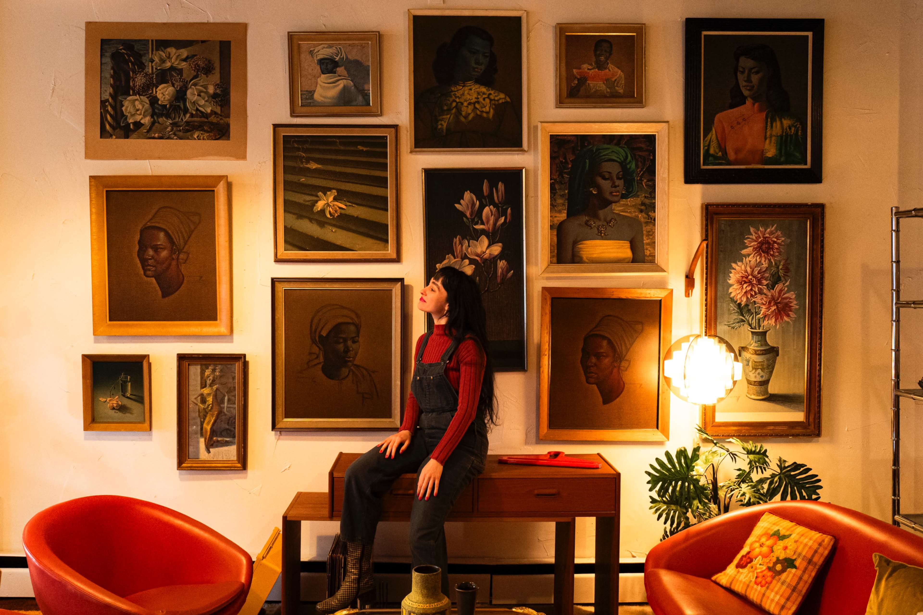 A woman sits on a wooden table in front of a gallery wall adorned with various framed portraits and artwork, with modern chairs nearby and warm lighting creating a cozy atmosphere.
