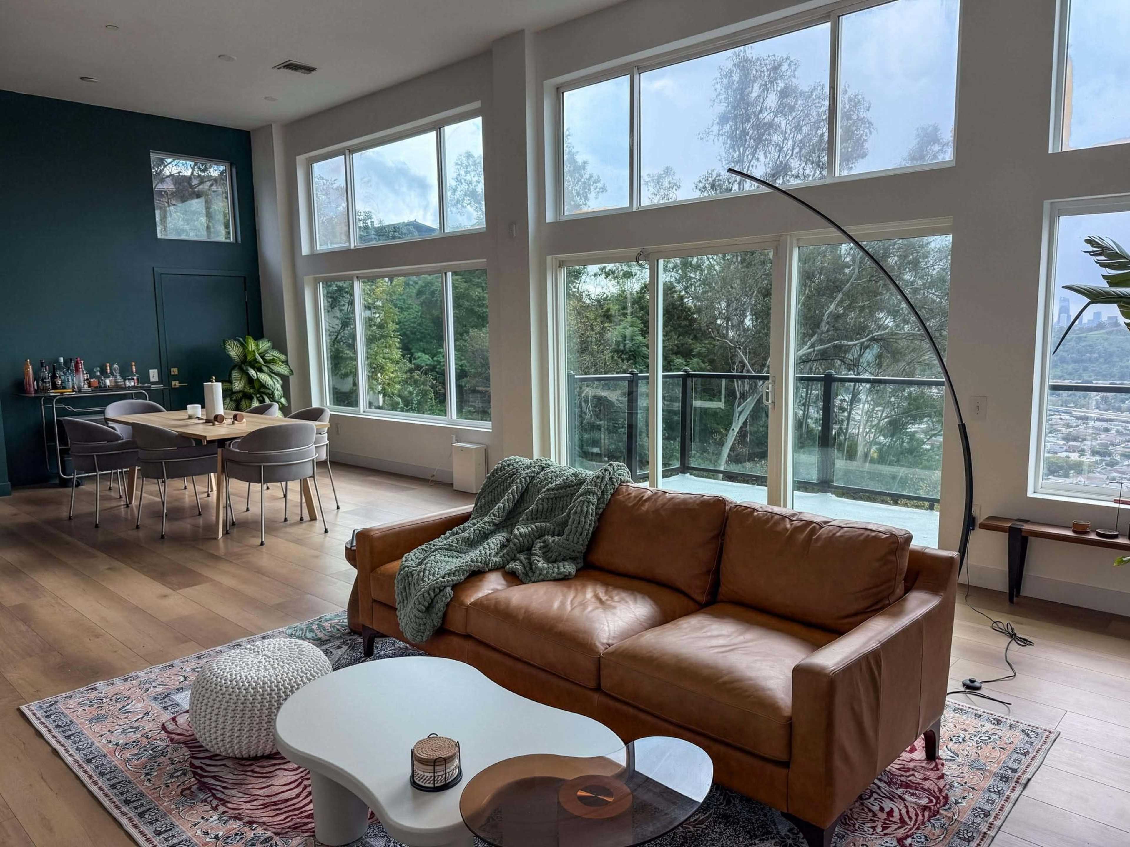 A living room features a brown leather sofa, a round coffee table, and large windows offering a view of trees and a hillside.