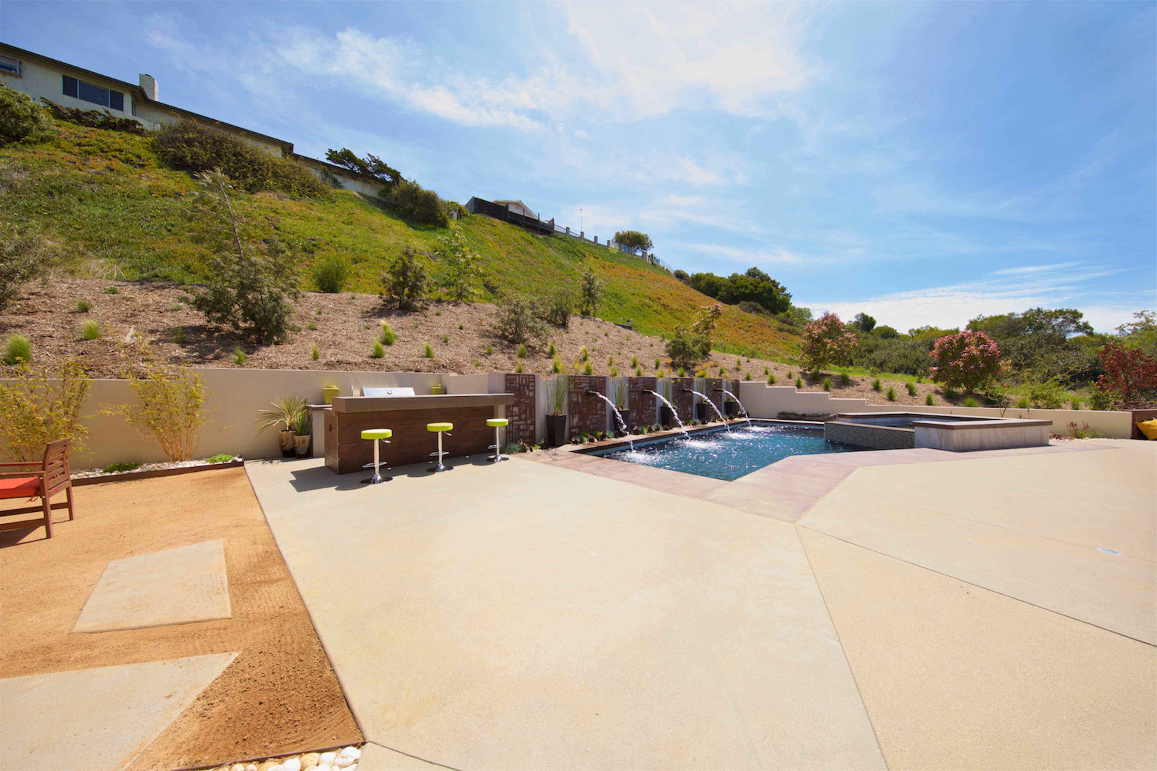 A modern outdoor pool area with a hot tub, bar seating, and a landscaped hillside in the background.