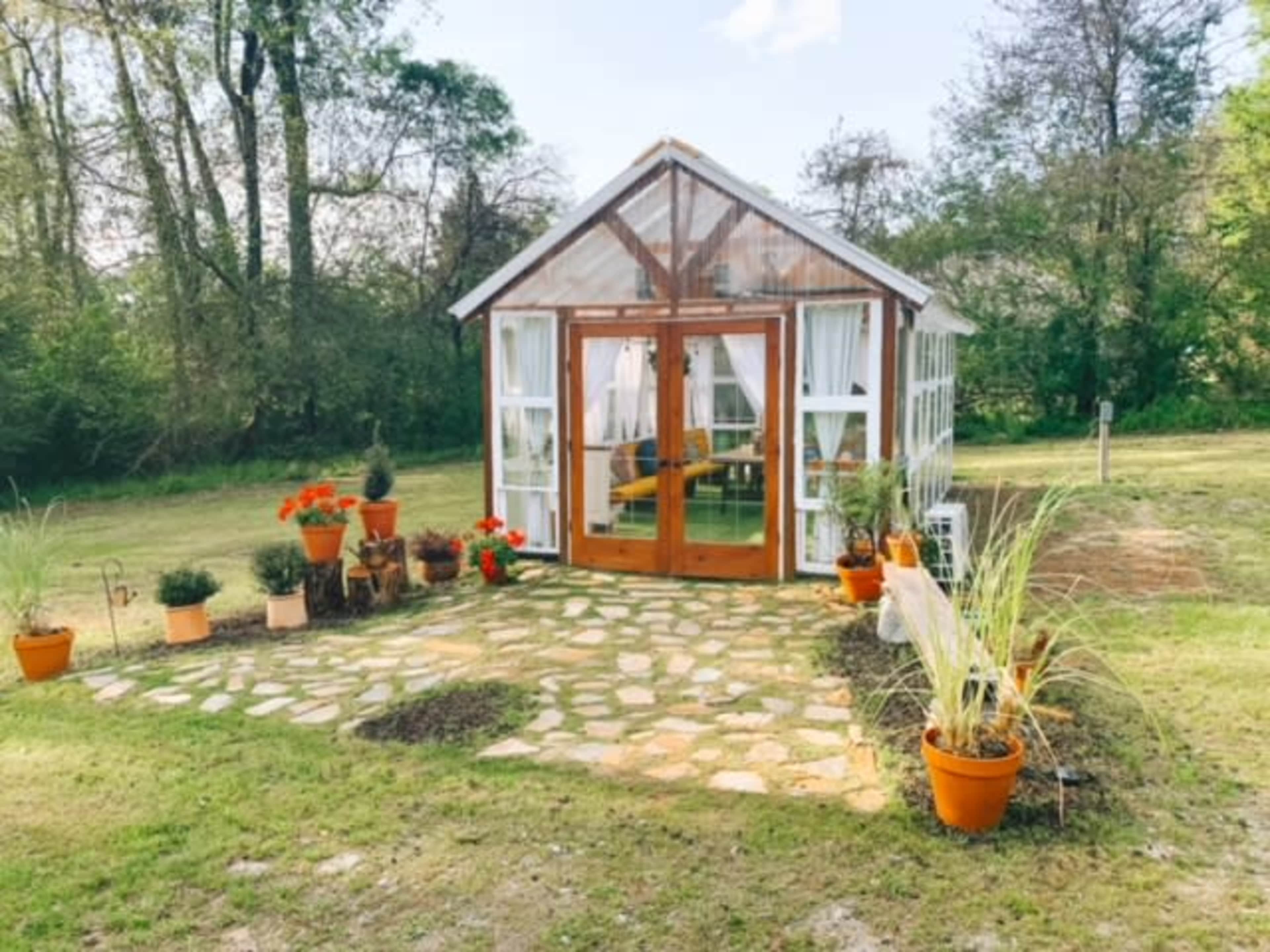 A glass greenhouse with wooden accents stands on a stone path surrounded by potted plants and flowers in a grassy area.
