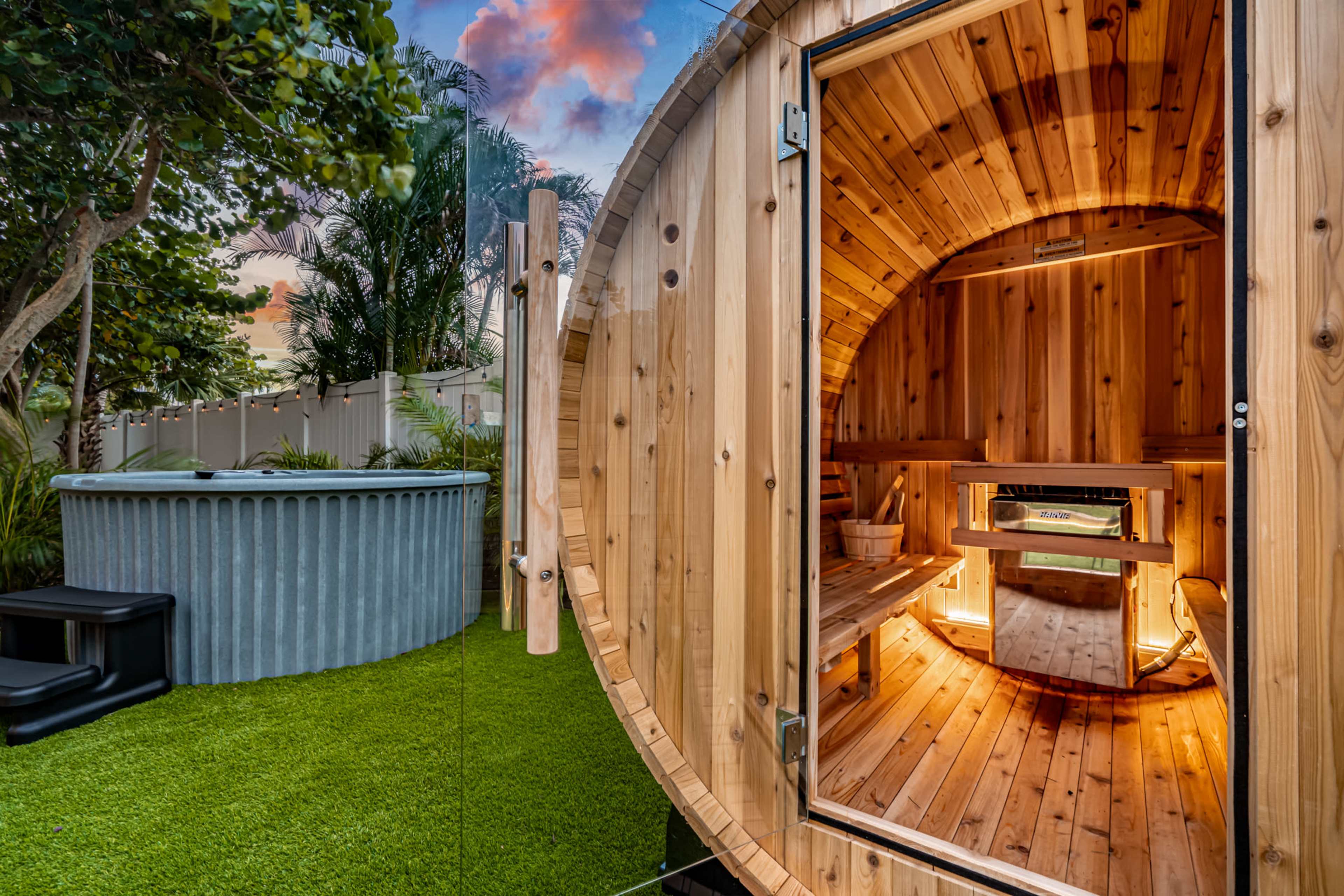 A wooden sauna with a circular design and an open door, adjacent to a round, galvanized metal hot tub in a grassy backyard setting.