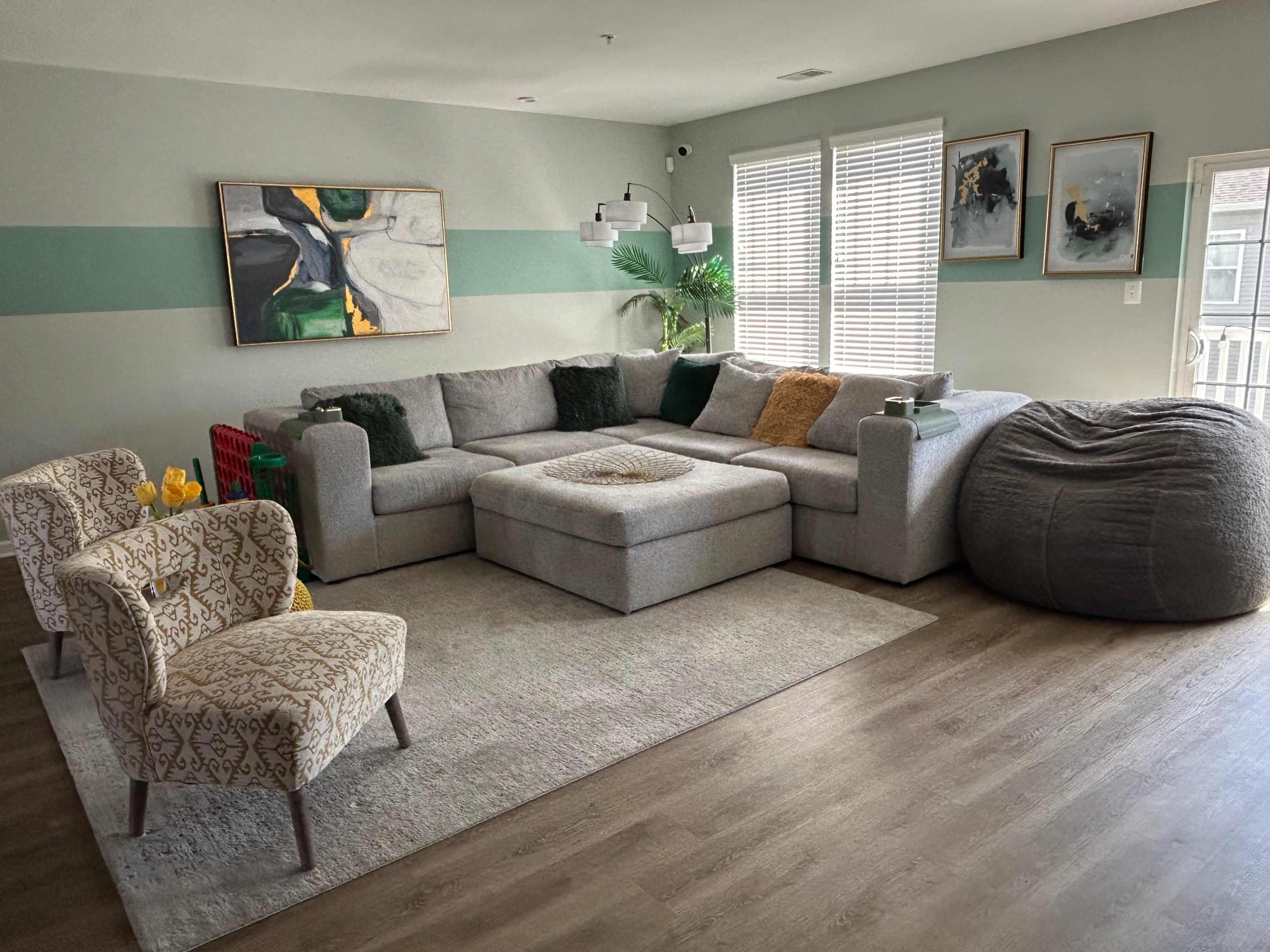 The image shows a modern living room with a large sectional sofa, a coffee table, a bean bag chair, and decorative armchairs, surrounded by green accent walls and large windows.