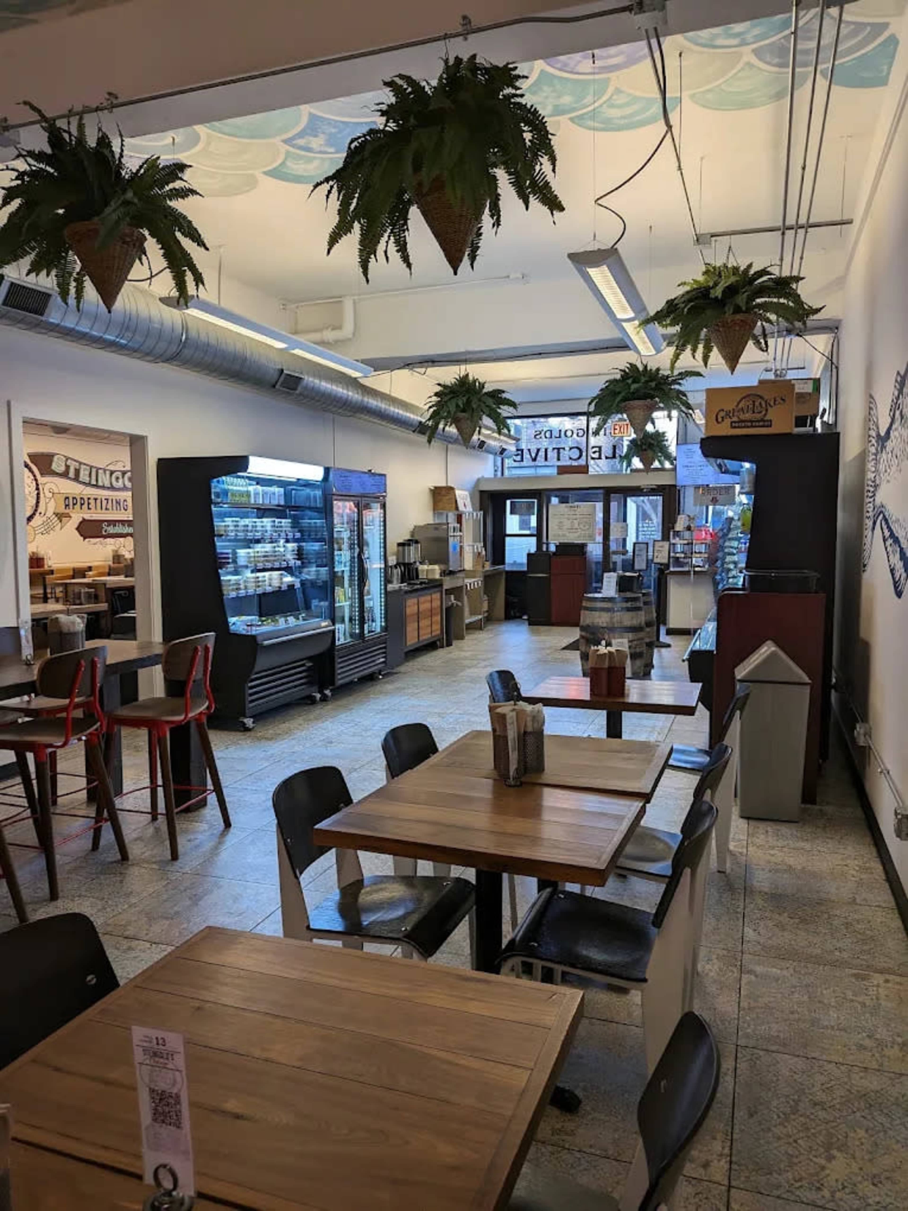 The image shows a brightly-lit cafe interior with wooden tables, chairs, and hanging plants, featuring a service counter and shelves with drinks.