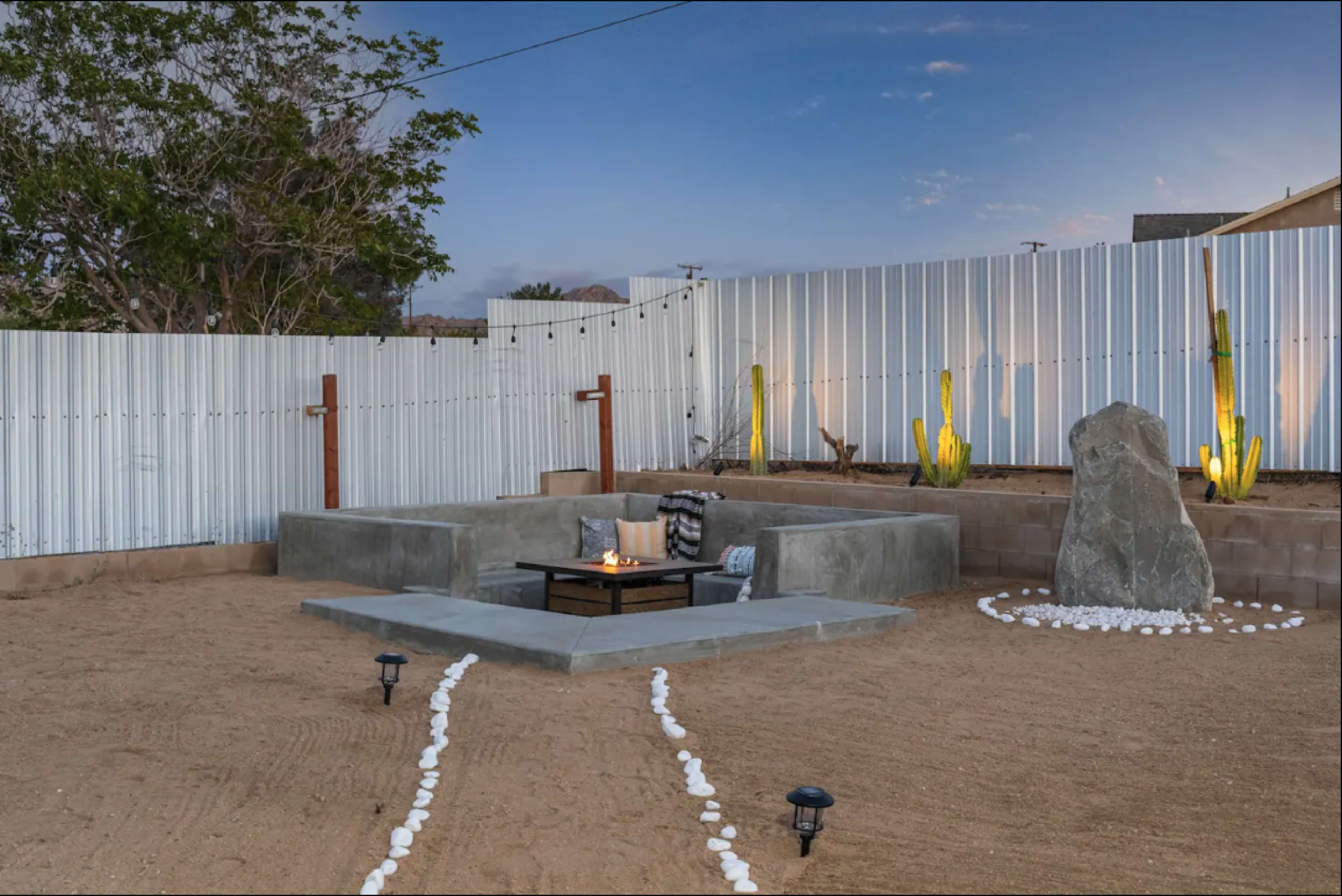 A minimalist backyard space with a raised seating area around a small fire pit, bordered by gravel and illuminated by outdoor lights.