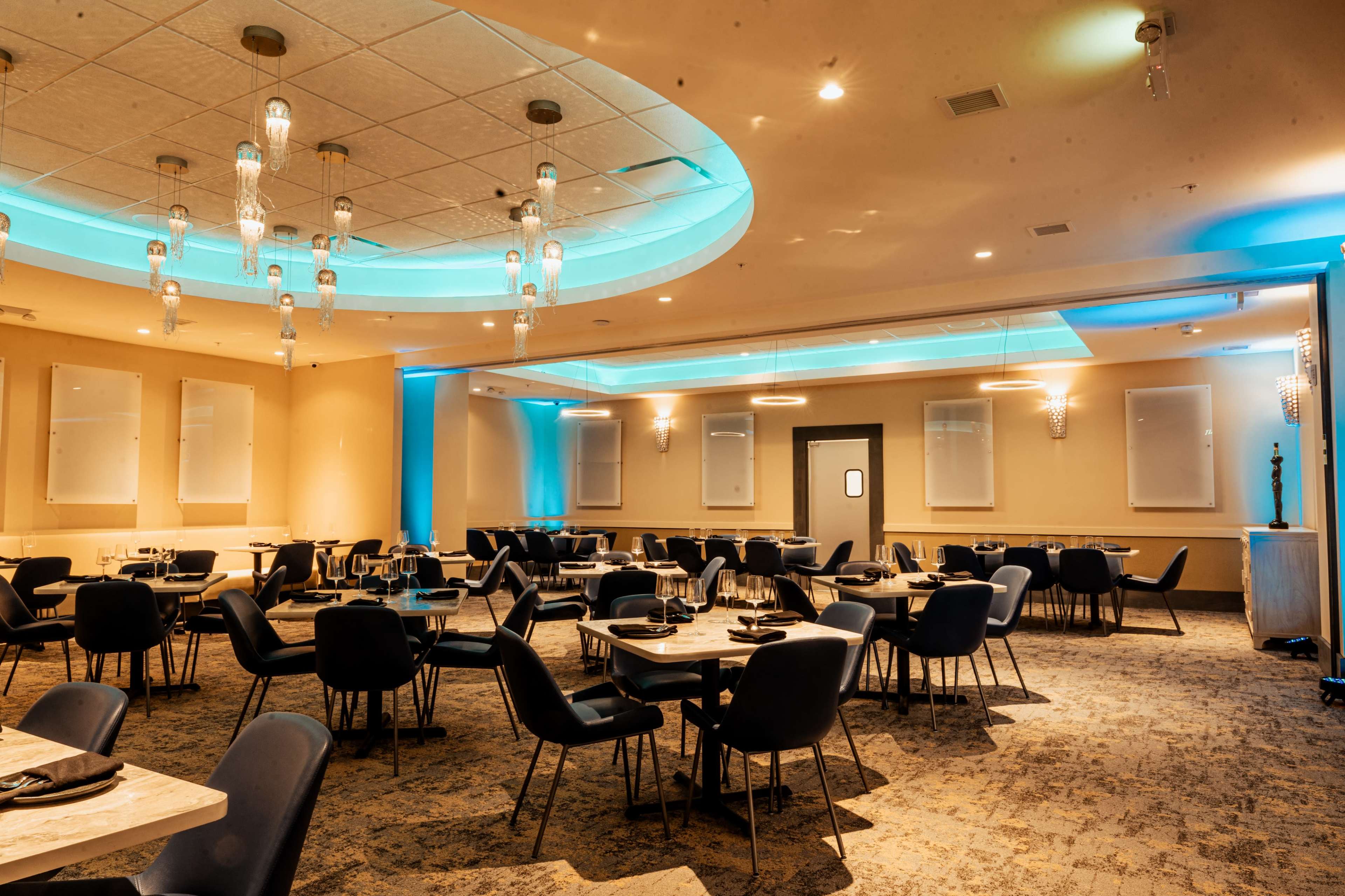 The image shows a modern restaurant interior with round tables set for dining, surrounded by blue accent lighting and decorative ceiling features.